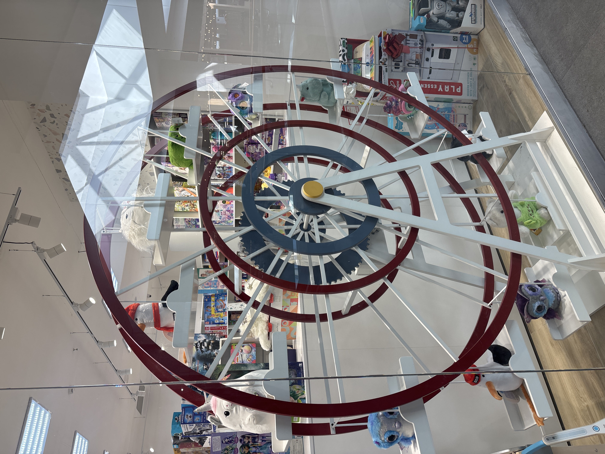 The image displays an interior retail store exhibit observed through a clear glass storefront in Izhevsk, Russia. The central feature is a stationary, large-scale Ferris wheel-style display unit. This structure is predominantly white, featuring red outer rings and a blue central gear-like component, with numerous white platforms serving as gondolas. Each platform holds a plush toy, including a white unicorn, a white penguin with a red hat and scarf, a blue multi-eyed furry creature, a green creature, and a purple owl. The background shows white shelving units stocked with various packaged children's merchandise, such as a "PLAY ESSENTIALS MODERN KITCHEN" set, a robot toy, and doll sets. The store interior is illuminated by overhead fluorescent light panels and track lighting. The flooring inside the store is a light brown wood-patterned surface, while the exterior foreground shows a grey tiled surface. No individuals or dynamic actions are visible within the frame.