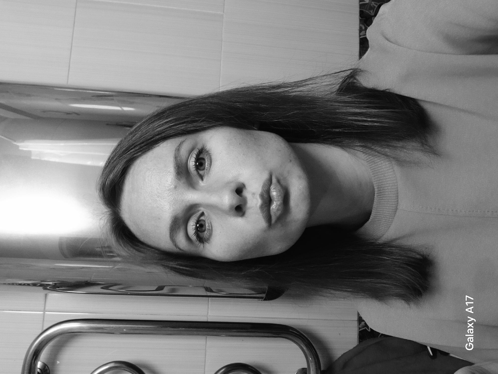 A monochrome photograph depicts a woman indoors. The location is likely a bathroom, indicated by tiled walls and a towel rack. The woman is positioned at an angle to the camera. Her hair is straight and medium length. She is wearing a crew neck sweater. The "Galaxy A17" watermark indicates the picture may have been taken by such a device. There is no evidence to indicate the location of Yekaterinburg, Russia in this photo.