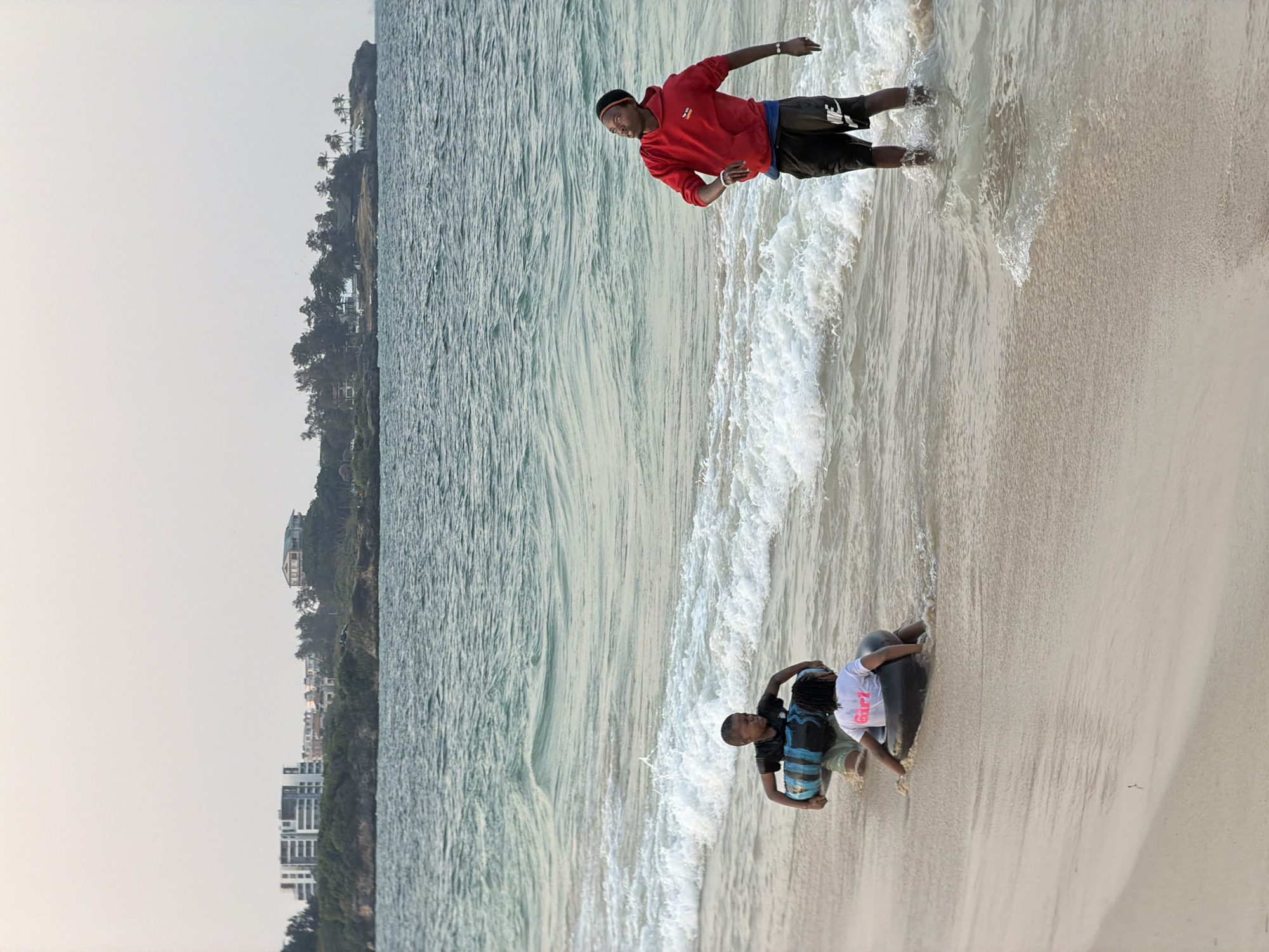 The image depicts a sandy beach in Dar es Salaam, Tanzania, with the Indian Ocean to the left. In the foreground, three individuals are interacting with the water. A person in a red hooded sweatshirt and black shorts stands in the shallow water, looking towards the right. To the left, two children are wading in the water. One child, wearing a black t-shirt and shorts, is carrying a large inner tube with blue and black stripes. Another child, wearing a white t-shirt with "Girl" printed on it, is seated within a dark inner tube. Gentle waves are washing ashore, creating a foamy surf. In the background, a low-lying coastline is visible, featuring a mix of trees and buildings, including what appear to be modern apartment complexes and houses. The sky is overcast and pale.