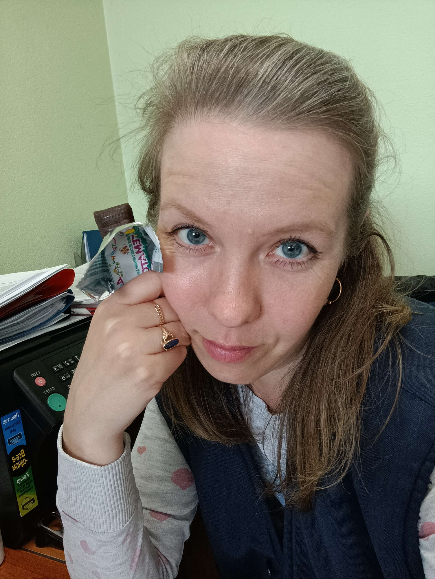 A fair-skinned woman with light brown hair and blue eyes is depicted in a close-up, selfie-style photograph. She is holding an opened, partially consumed tube of "Lew Tome" (a product name) next to her left eye, cradling her head with her hand. The ring on her finger is gold and features a dark-colored stone. She appears to be indoors, possibly in an office setting. In the background, a multifunction printer is visible with a sticker in Russian. A stack of papers is on the printer. The Russian sticker on the printer includes information, like "BCE-B" and "MONDO." Given the "Lew Tome" product and the Russian sticker, it can be inferred the picture was taken in a Russian-speaking location, possibly Chaikovsky, Russia.