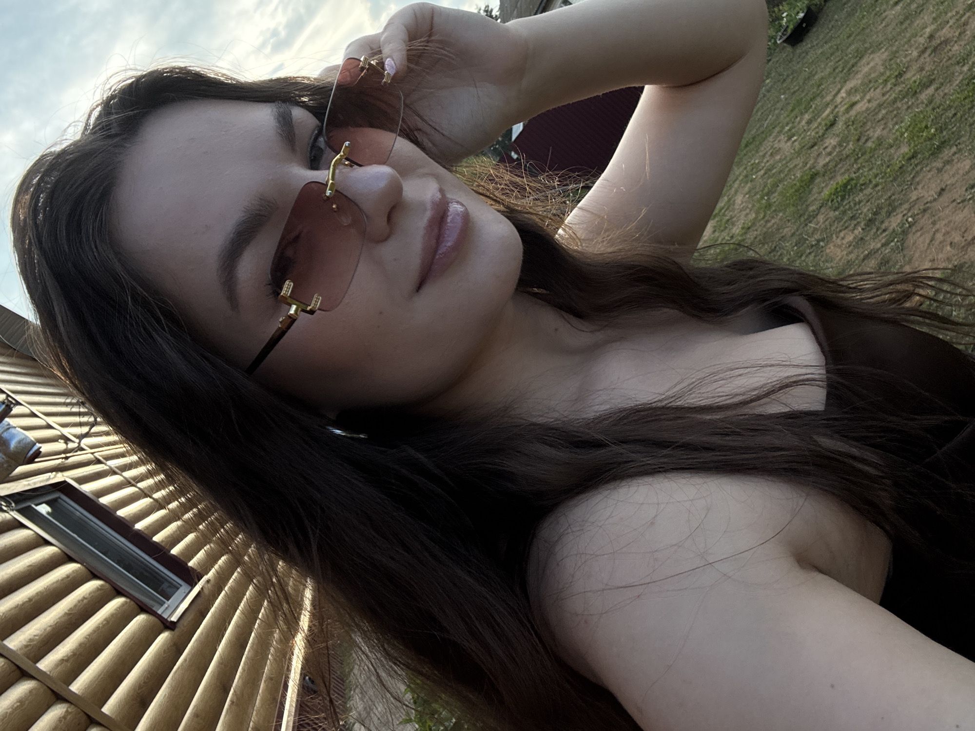 In Rusalevka, Russia, a young woman with long, dark brown hair and fair skin is captured in a close-up, outdoor photograph. She is wearing a dark brown top and stylish rimless sunglasses with pink lenses, which she is adjusting with her right hand. Her left shoulder and part of her chest are visible. The background features a wooden structure, possibly a house, with horizontal log siding and a small window. Beyond that, a green grassy area with some small white flowers and a dark circular object can be seen, under a bright, slightly cloudy sky. The lighting suggests it is daytime, possibly late afternoon or early evening, with sunlight casting shadows on the wooden wall.