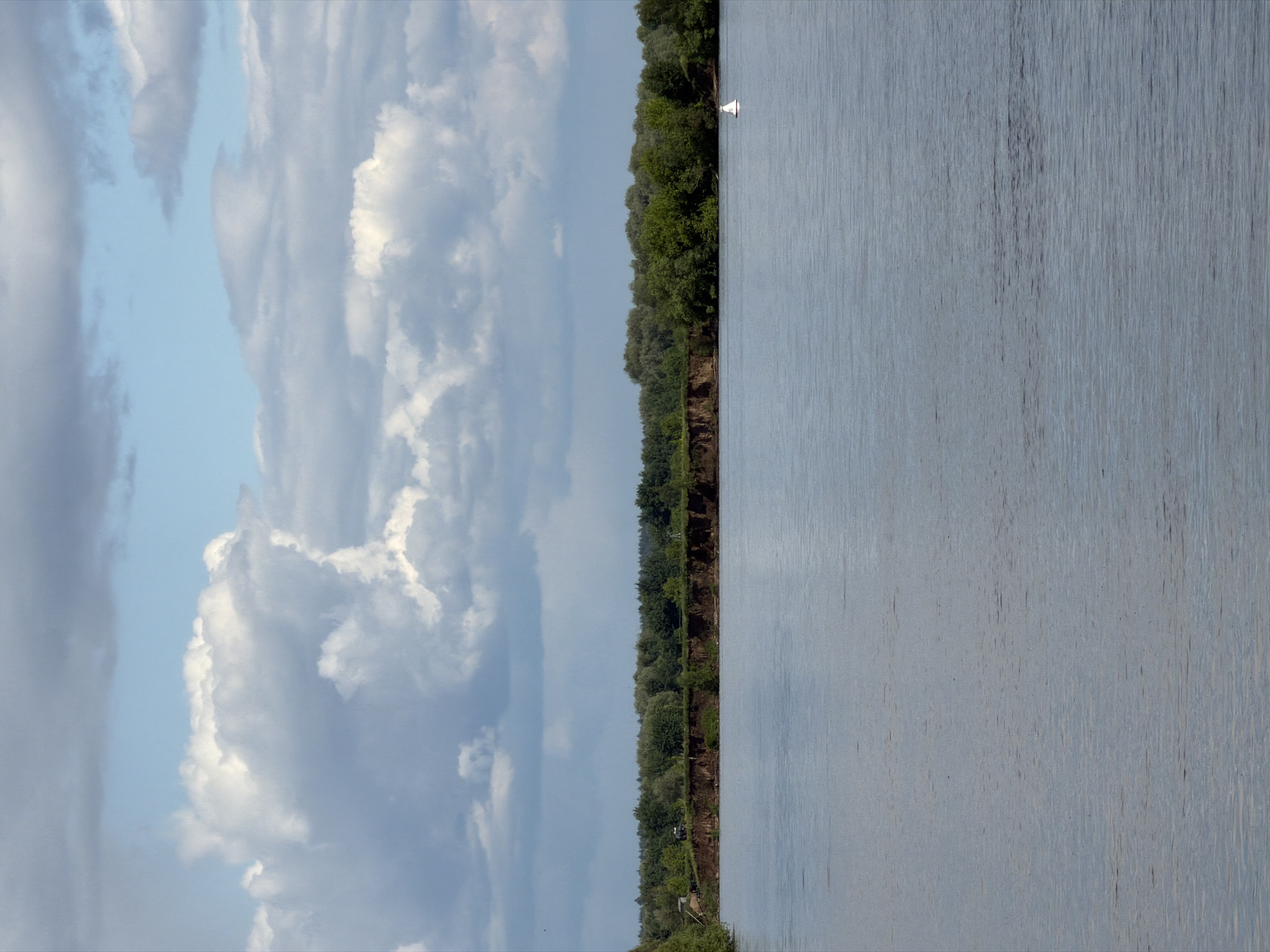 The image shows a body of water, possibly a river, with a shoreline covered in trees. The sky is filled with cumulus clouds under a blue sky. The perspective suggests the photo was taken from a low angle, perhaps near the water level. There is a single white object on the water that appears to be floating. The image lacks people or any definitive markers to confirm it is specifically Zaozerye, Russia. The scene is a natural waterscape with sky, water, and vegetation as the primary elements.