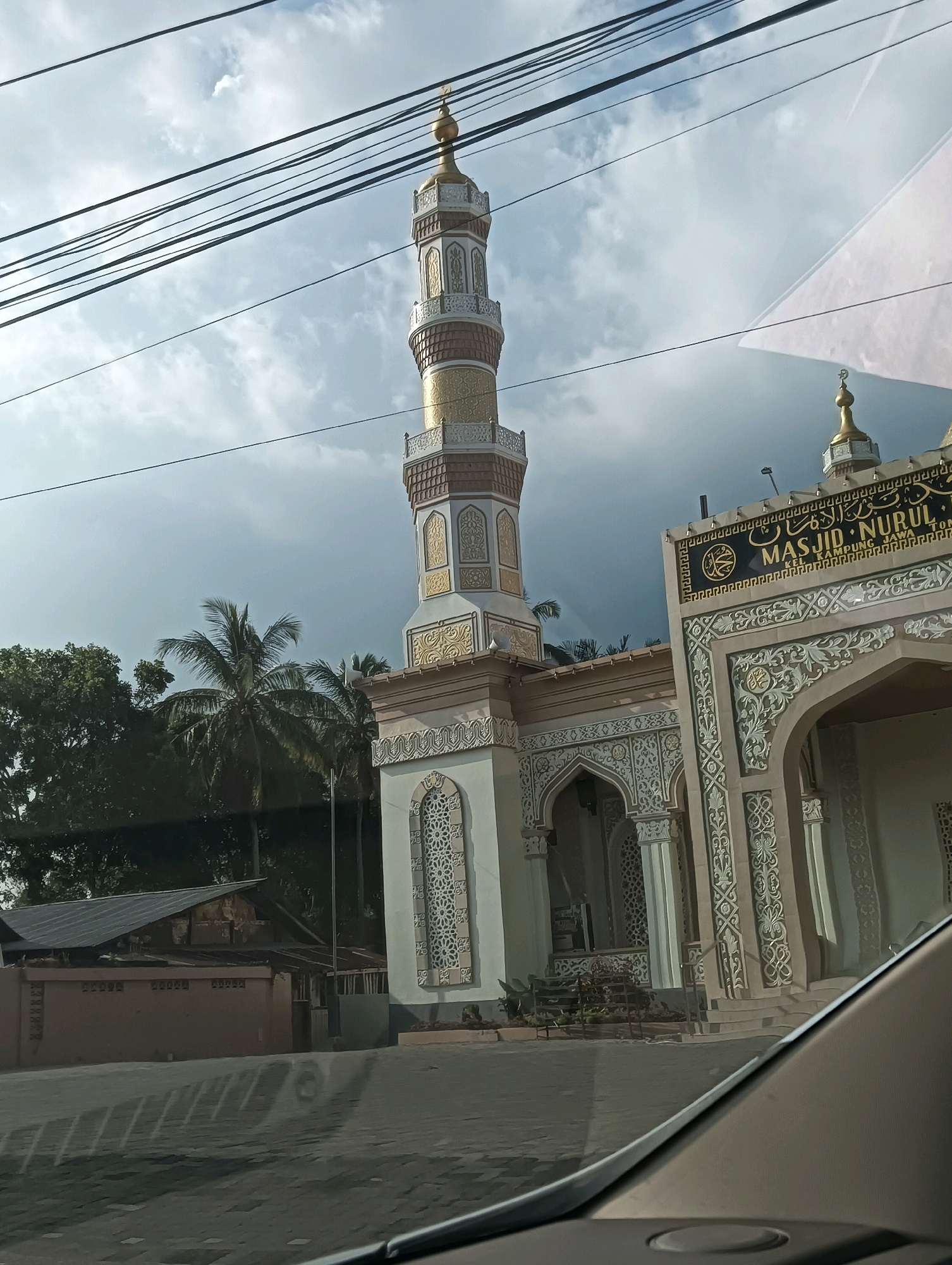 The image shows the exterior of the Masjid Nurul, located in Tomohon, Indonesia. The mosque features a prominent minaret with a golden finial, intricate decorative patterns on its facade, and an arched entrance. The mosque is situated in an area with trees, including a palm tree, and a building with a corrugated metal roof is visible to the left. Power lines stretch across the sky, obscuring parts of the blue, cloudy sky. The foreground shows a paved surface, possibly a road, with shadows cast upon it. The reflection of the sky and clouds is visible on what appears to be a car windshield, suggesting the photo was taken from inside a vehicle. There are no people visible in the image.