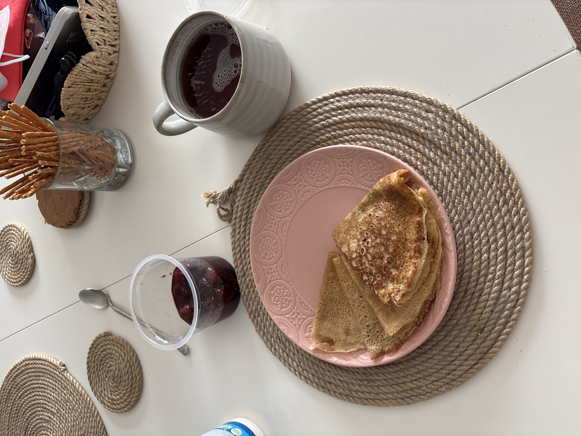 The image depicts a table setting with a meal. A large mug filled with a dark liquid sits near a glass containing breadsticks. A plastic cup containing berries and a spoon are also present. A pink plate on a woven placemat holds a stack of crepes. The setting includes a variety of round, woven coasters. A woven basket can be seen in the upper left corner. A blue and white container is partially visible near the bottom center of the image. The scene suggests a breakfast or brunch setting. There is no verifiable information suggesting that this is Krasnoyarsk, Russia.