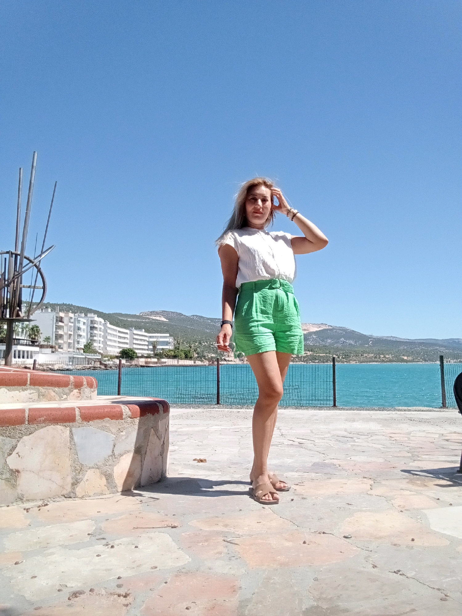 A fair-skinned woman with blonde hair and a white top and green shorts poses for a photo outdoors. She is standing on a stone pathway with a body of water and buildings in the background. She has her right hand on her head. The scene appears to be a sunny day with a clear blue sky. The location is the Silifke city in Turkey.