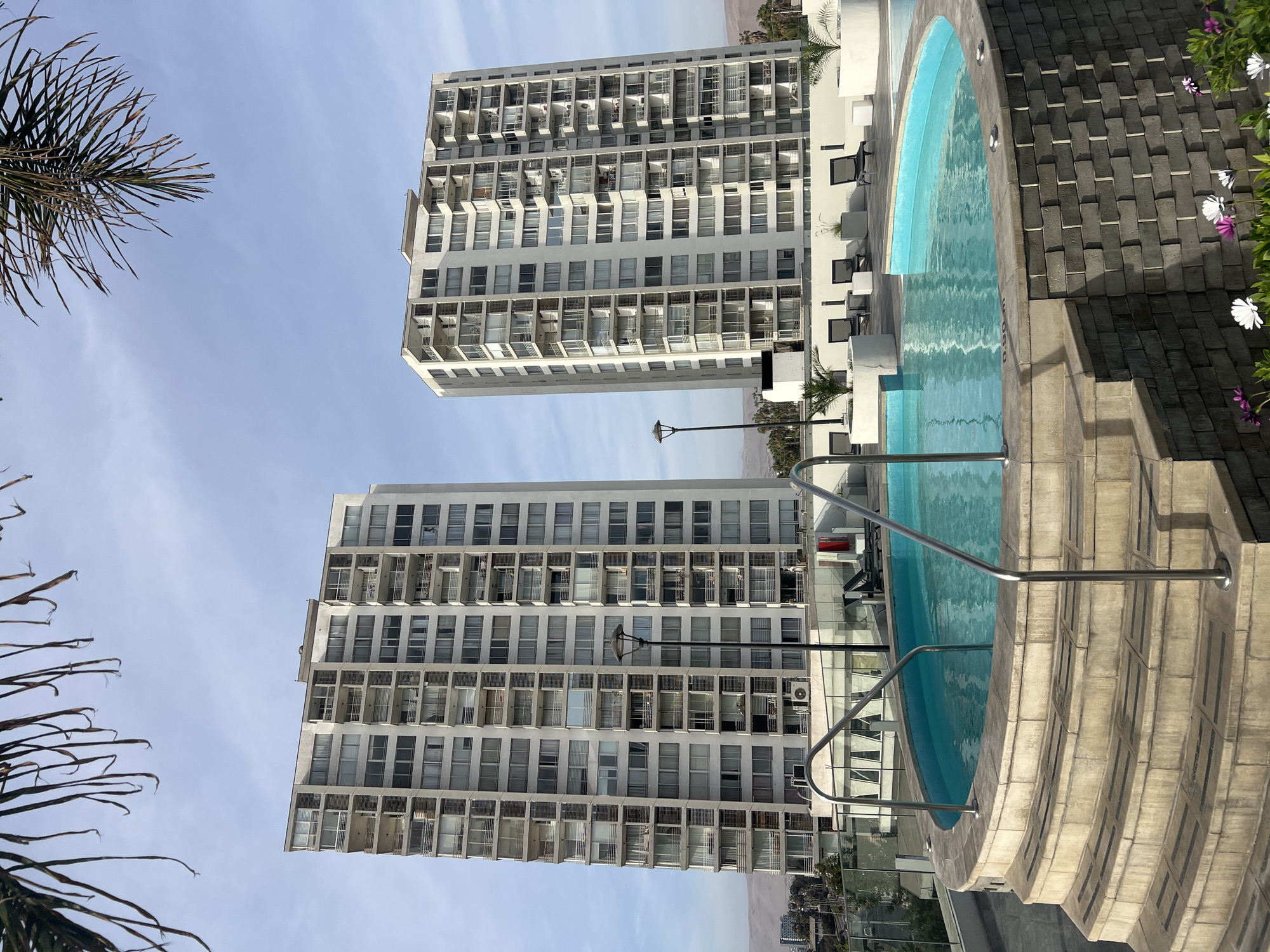 The image depicts a high-angle view of a modern apartment complex and swimming pool area located in Arica, Chile. Two multi-story apartment buildings with numerous balconies are visible against a clear blue sky. A circular swimming pool with blue water and a stainless steel railing is in the foreground. The pool is surrounded by a tiled deck. A palm tree branch appears in the corner of the image, adding a tropical element to the scene. The city and desert landscape are visible in the background. There are no visible people in the picture.