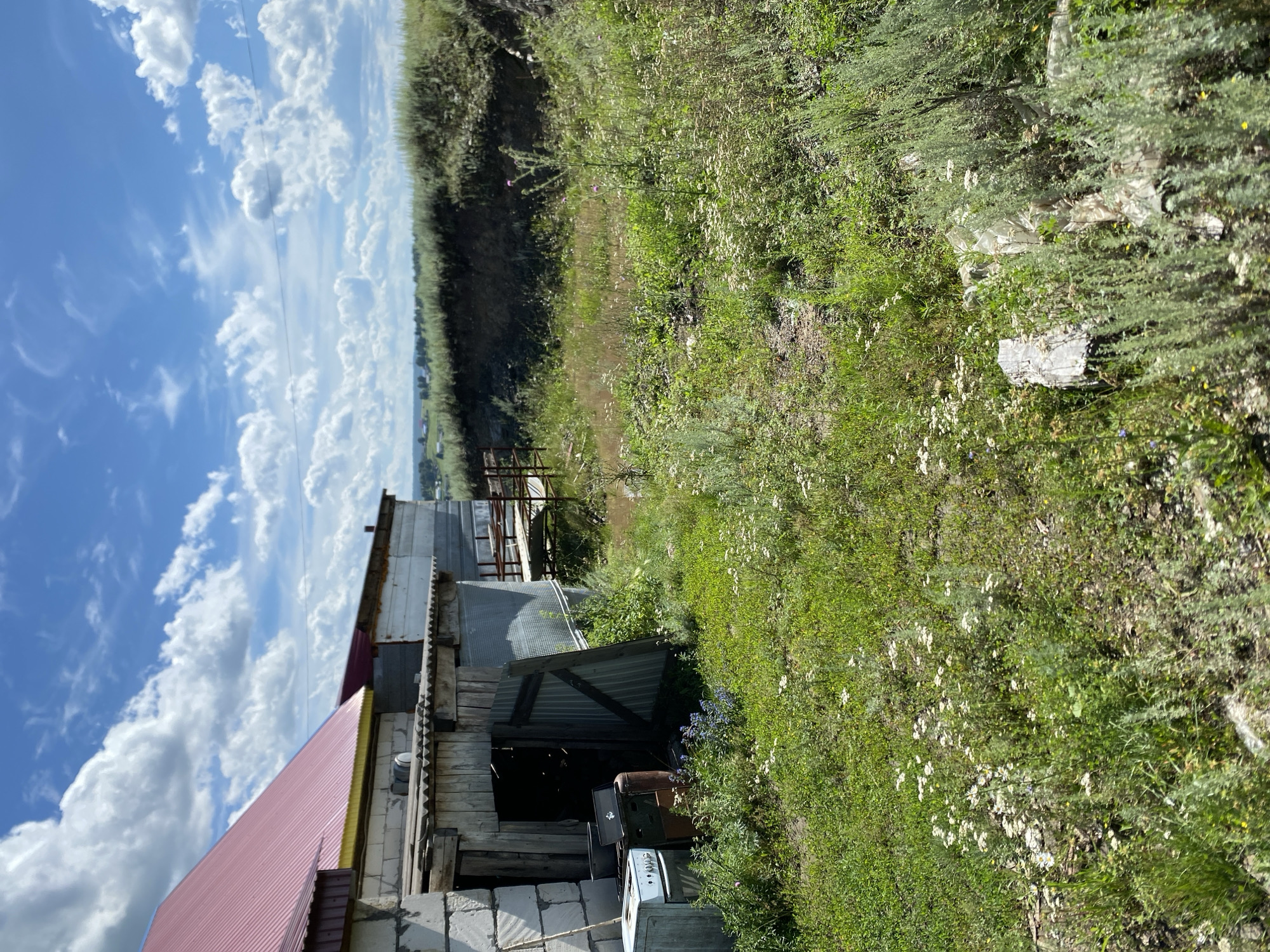 The image depicts an outdoor scene in Yuldybayevo, Russia, featuring an unkempt yard with various structures and vegetation. In the foreground and mid-ground, lush green grass and wild flowers dominate the landscape, with patches of dry earth and scattered debris. To the left, a dilapidated building with a red metal roof and concrete block walls is visible. Adjacent to this building is a lean-to structure with a wooden and corrugated metal facade. Outside this lean-to, an old white electric stove and rusty metal containers are placed. Further to the right, a slope covered in dense green brush descends towards a distant horizon where a few scattered houses and trees can be observed. The sky is a vibrant blue, adorned with fluffy white clouds, indicative of a clear, sunny day. There are no people or animals present in the scene.