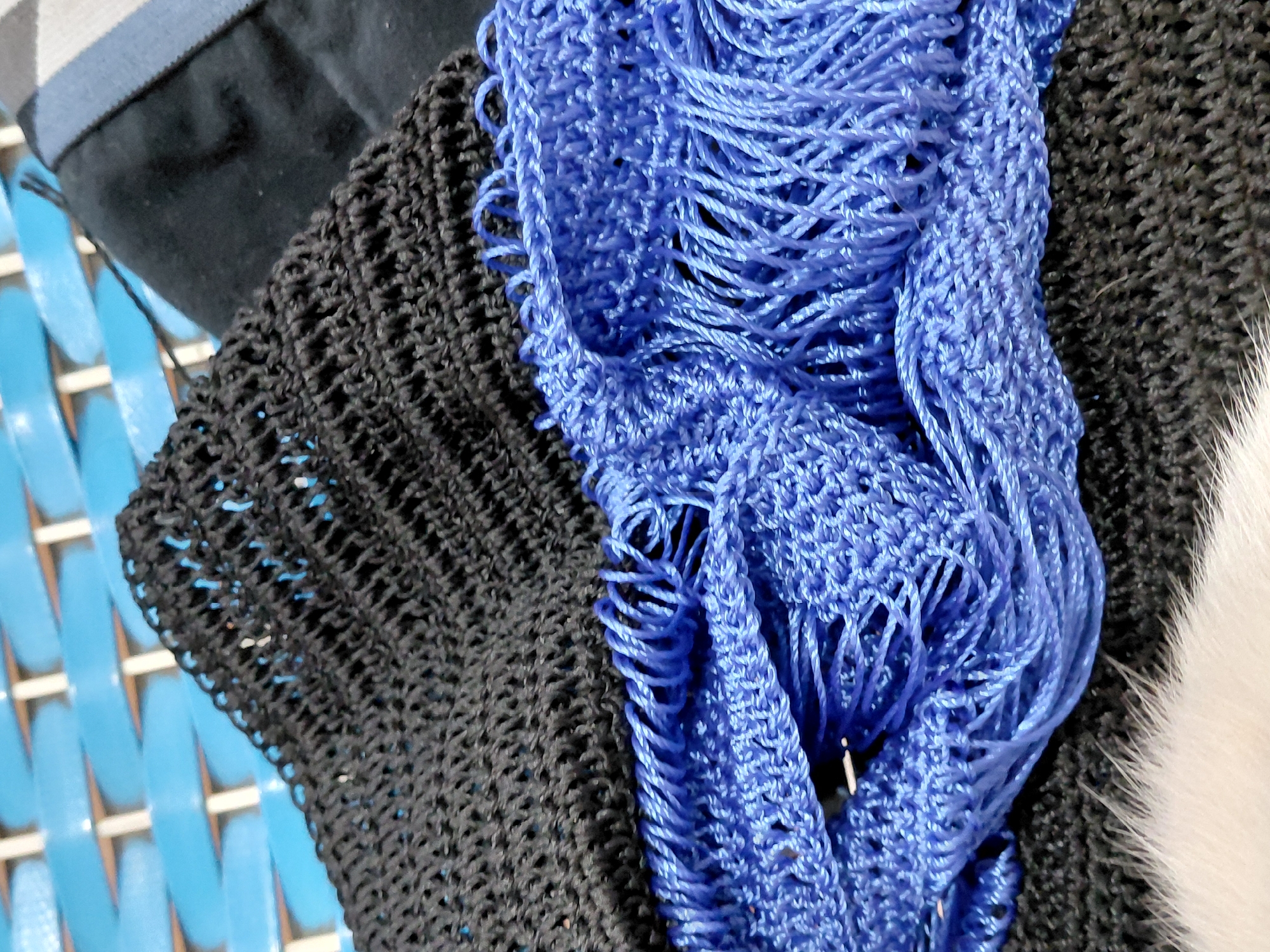 The image shows a close-up of several fabric items. A black, knitted garment is visible on the left and right sides. A bright blue, loosely knitted scarf or wrap with fringed edges is draped over the black fabric. The items appear to be resting on a blue and white slatted surface. The location is unspecified.