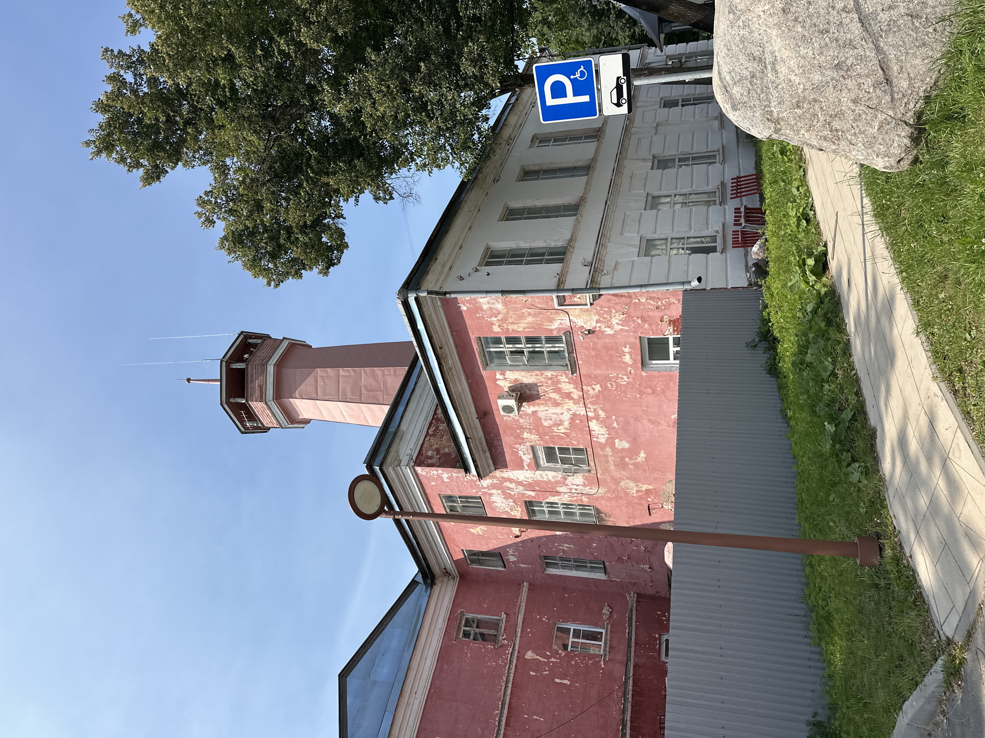 The image shows a red and white building with a pink tower in Selivanovo, Russia, on a sunny day. The building appears to be old, with peeling paint on the red sections. There are several windows visible, some with glass and others broken. A gray corrugated metal fence runs in front of the building, separating it from a grassy area and a sidewalk. A brown lamppost stands on the sidewalk. To the right of the building, there is a blue sign with a white "P" and a wheelchair symbol, indicating disabled parking. Next to that is another sign with a white car silhouette on a black background. A large tree with green foliage is visible behind the building and sign. In the foreground, a large grey rock sits on the grass next to the sidewalk.