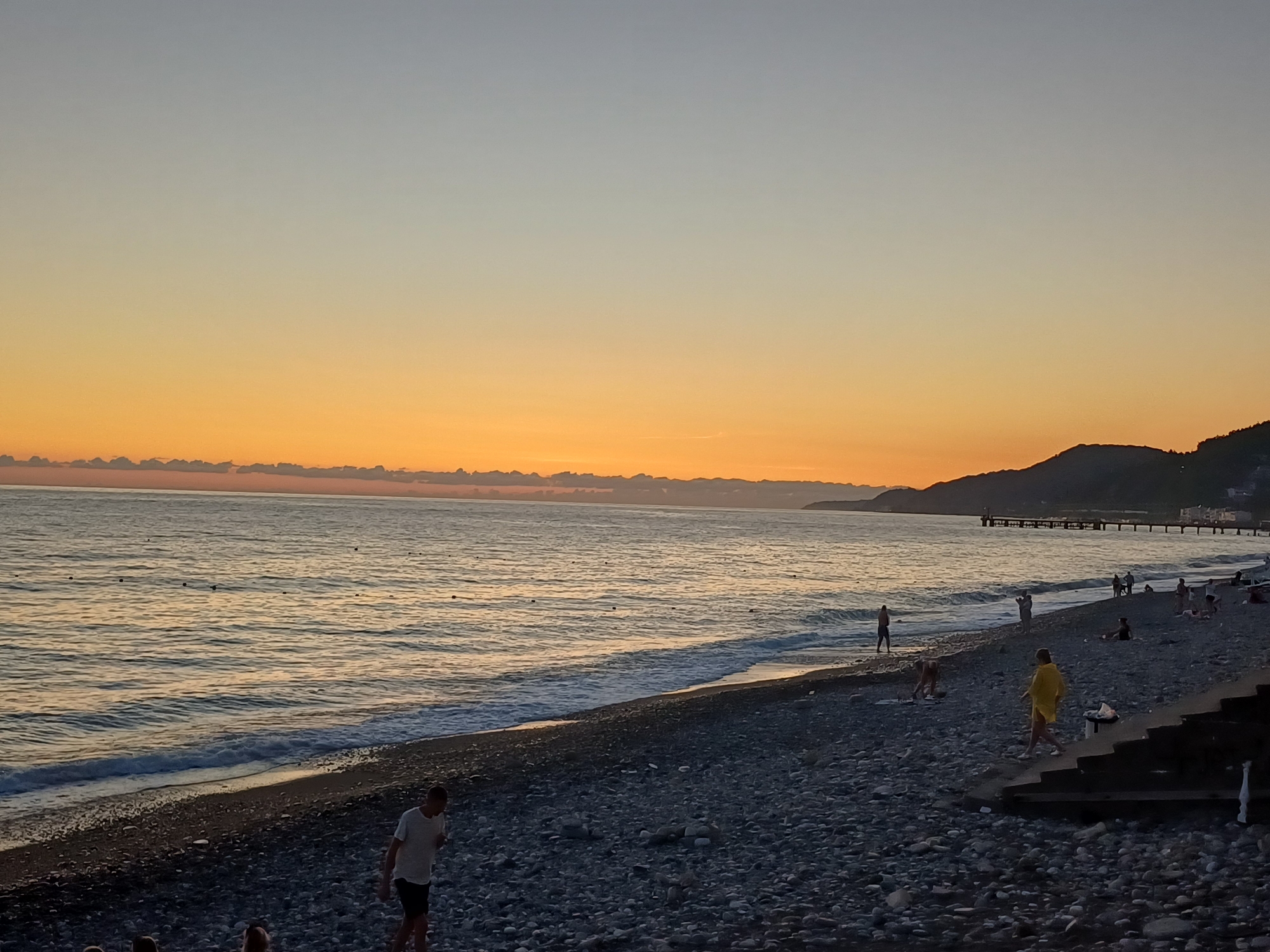 A wide shot captures a pebble beach in Sochi, Russia, at dusk. The sky is a gradient of pale blue to warm orange near the horizon, with a few clouds visible. The sea is calm, with gentle waves lapping the shore. The beach is populated with a few people. In the foreground, a man in a white t-shirt and black shorts walks on the pebbles. Further down the beach, a person in a yellow garment walks towards a set of dark stairs. Other individuals are scattered along the shoreline, some standing and others sitting. In the distance, a pier extends into the sea, and a dark, hilly coastline is visible. The overall scene depicts a peaceful evening at the seaside.