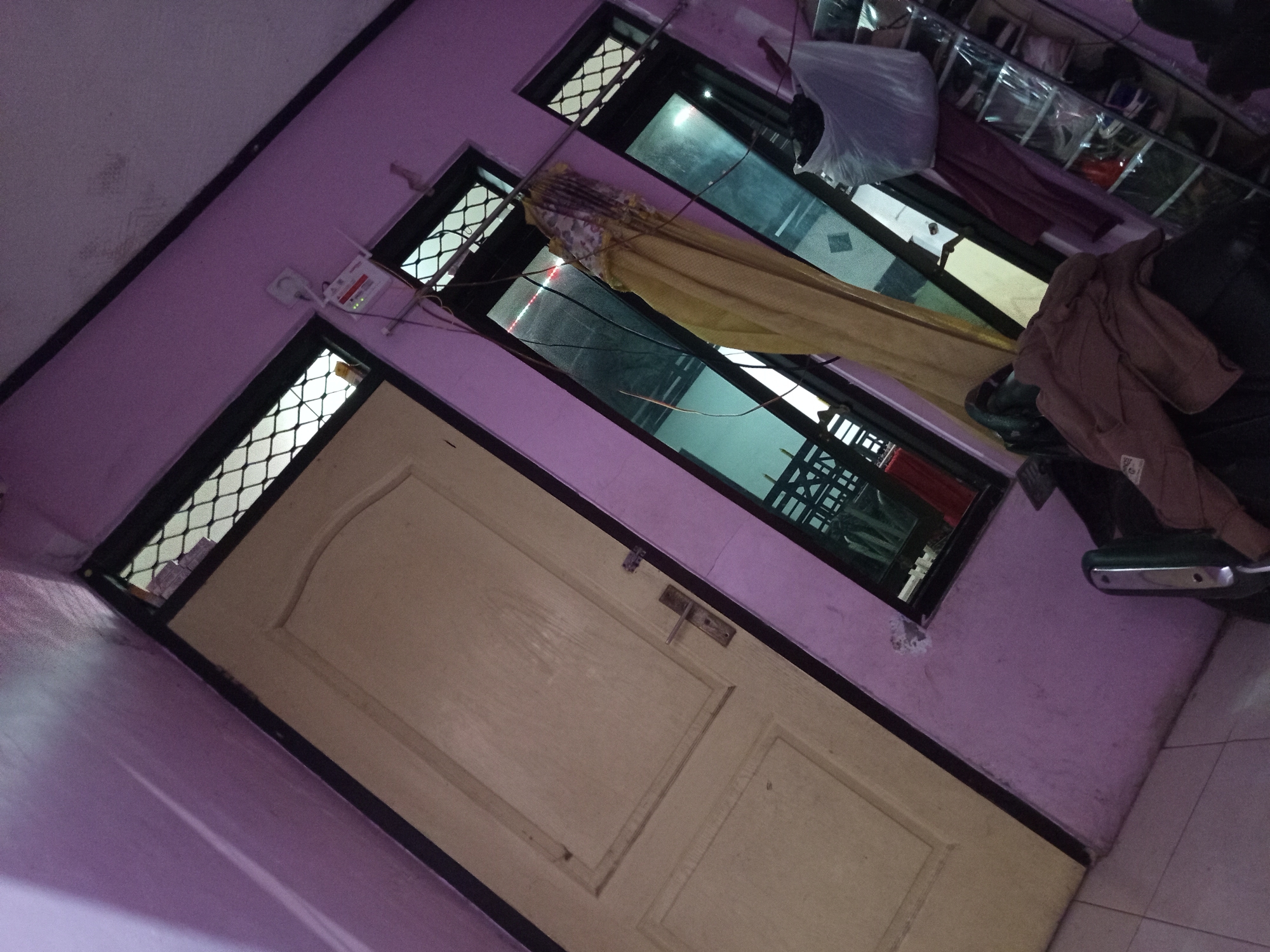 The image depicts an interior scene, likely a residential hallway or room, with a strong purple hue on the walls. A light-colored door with paneled detailing is visible on the left side of the frame, featuring a metallic door handle. To the right of the door are two tall, narrow windows with a crisscross pattern on the upper portion, suggesting security bars or decorative grating. The windows reflect a dimly lit interior.
A yellow curtain or fabric is draped across the space between the two windows. There are numerous wires and cables hanging from the ceiling, some of which are connected to a white Wi-Fi router mounted on the wall above the right window. Next to the right window, a clear plastic shoe organizer filled with shoes is mounted on the wall. A crumpled white plastic bag and dark-colored clothing items are placed on top of the shoe organizer and a dark-colored piece of furniture in the foreground.
The image is taken at an angle, giving a sense of the room's proportions. The floor appears to be tiled. There are no people visible in the image. Based on the reference to the city of Tebel, Indonesia, this scene is located within that region.