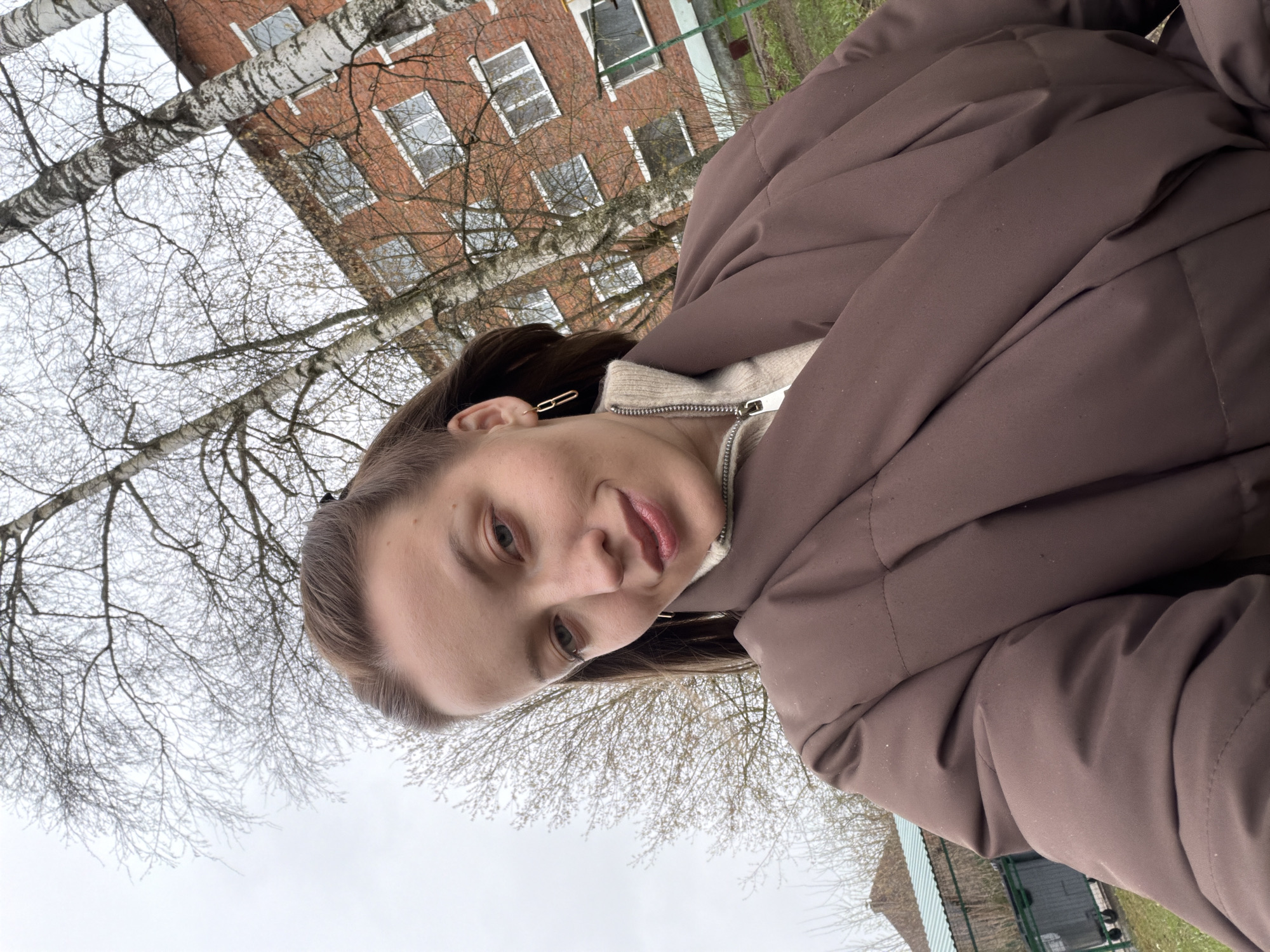 The image is a portrait-oriented photograph rotated 90 degrees counter-clockwise, capturing a young woman in an outdoor setting. The woman, positioned on the right side of the frame, has brown hair and looks towards the left with a slight, gentle smile. She is wearing a dark brown puffy jacket over a light-colored, possibly cream or beige, turtleneck sweater. A distinct golden chain-link earring is visible in her left ear.
The background features a grey, overcast sky. Numerous bare branches of birch trees, characterized by their white bark, crisscross the upper and central parts of the frame, suggesting a late autumn, winter, or early spring season. Behind the trees, a multi-story brick building with rectangular windows is visible in the upper left quadrant of the image. A small patch of green grass or foliage can be seen in the far upper right corner.
The scene depicts a calm daytime moment, likely in a residential or urban area, under a cool or cold weather condition, given the attire and bare trees. The presence of birch trees and the style of the brick building are consistent with the context of Reshetnikovo, Russia. There is no visible text in the image.