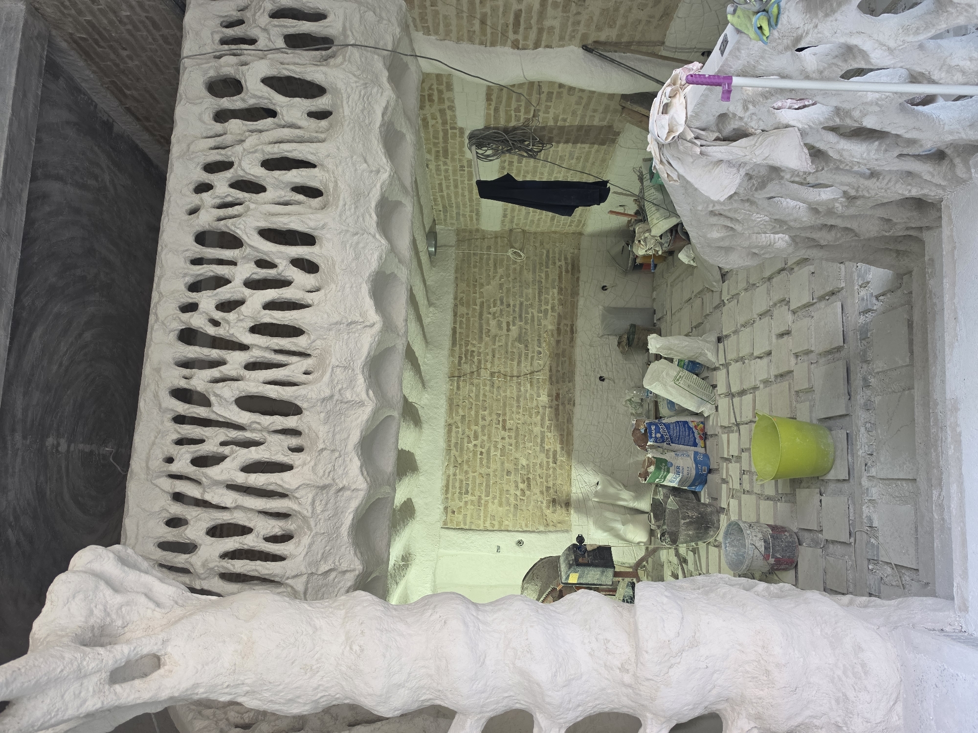 This image shows the interior of a room under construction or renovation. The dominant features are decorative, organic-shaped structures made of a white, textured material, resembling natural formations like coral or bone. These structures form a railing or balustrade and a column. The room has exposed brick walls, a concrete ceiling with swirling patterns, and a floor covered in square tiles, some of which appear to be missing or unfinished. Various construction materials are visible, including bags of cement or plaster, buckets, a cement mixer, and tools. A dark garment hangs on a hook on the wall. The scene suggests an ongoing project, possibly an artistic or architectural endeavor, given the unconventional design elements. The location is identified as Varna, Bulgaria, although no specific city landmarks are visible to confirm this.