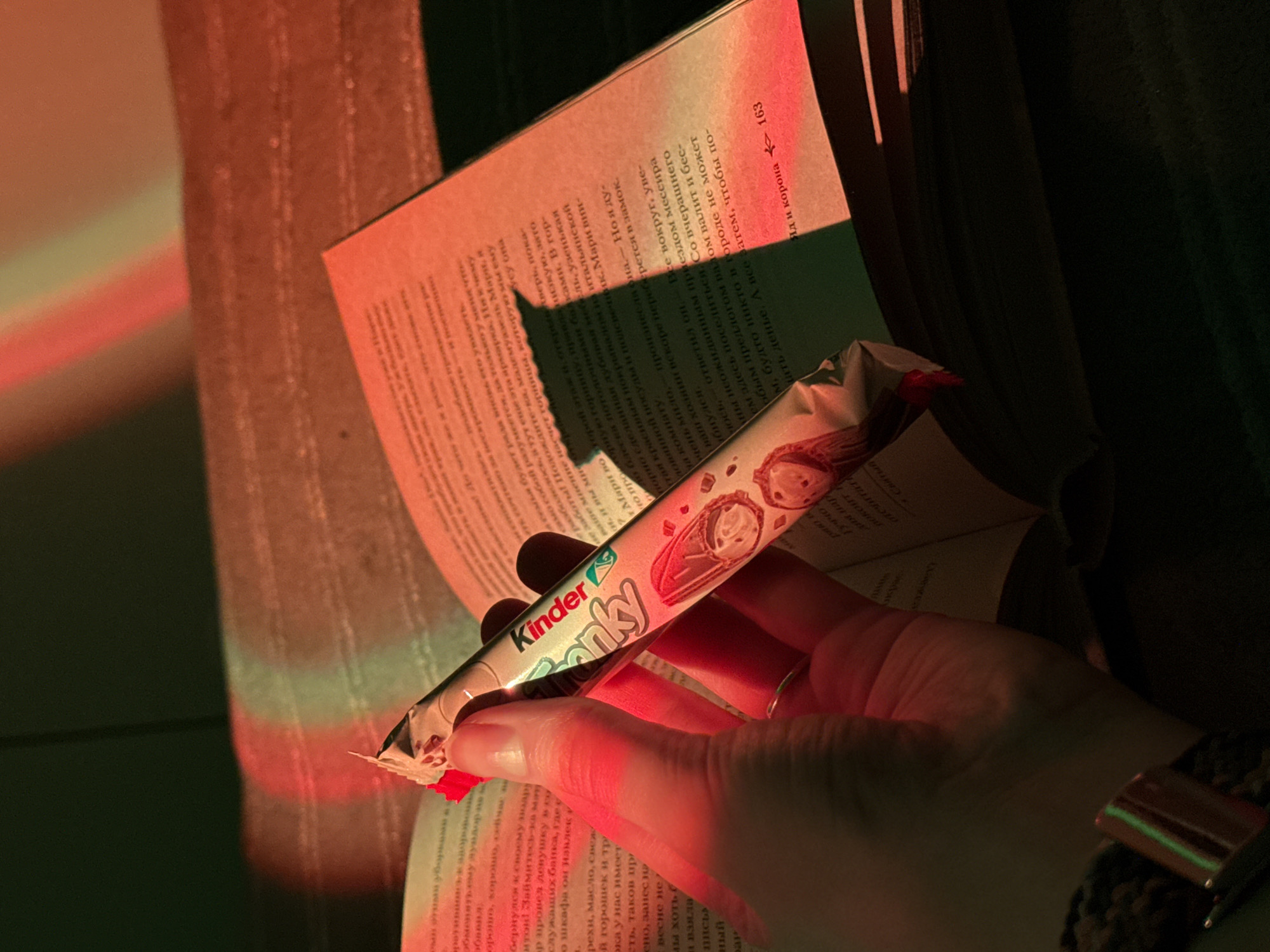 A person holds a Kinder Bueno bar while reading a book. The book is open to a page with Russian text. The lighting is red and green. The person is wearing a watch.