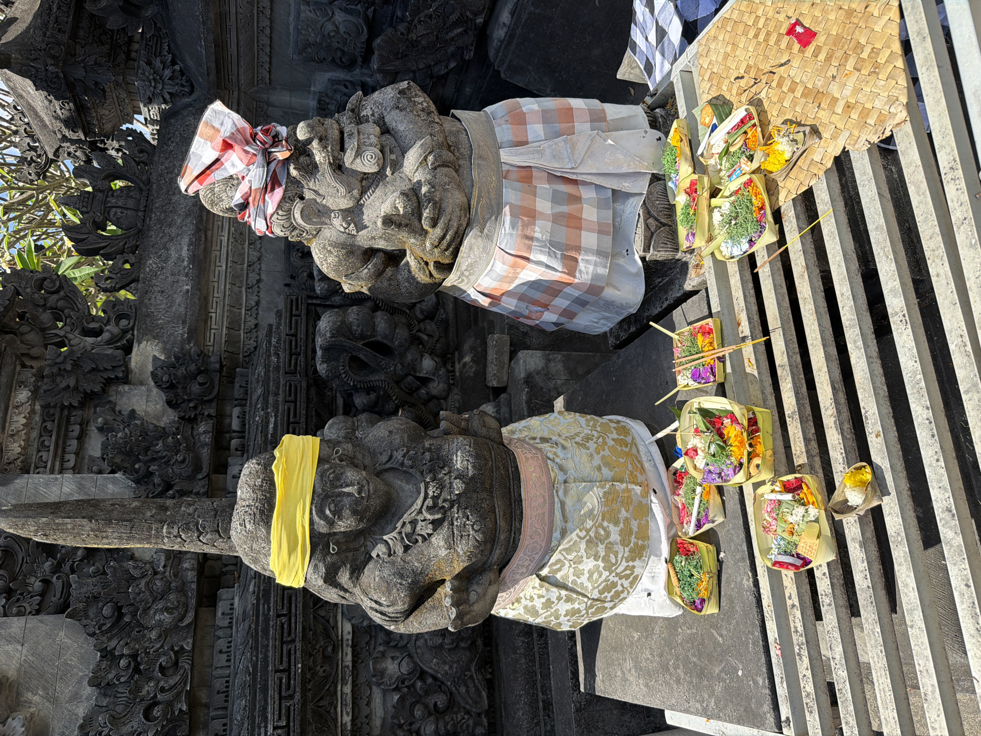 Two stone statues, likely temple guardians, stand on a stone platform in front of a stone temple structure in Kuta, Indonesia. The statue on the left has a yellow fabric band around its head and wears a patterned cloth skirt. The statue on the right has a red and white checkered fabric band around its head and wears a similar patterned cloth skirt. In front of the statues are several small woven baskets filled with offerings of colorful flowers, leaves, rice, and incense sticks. The offerings are arranged on a metal grate and a woven mat. The environment appears to be outdoors, with sunlight casting shadows. The temple structure in the background features intricate stone carvings.
