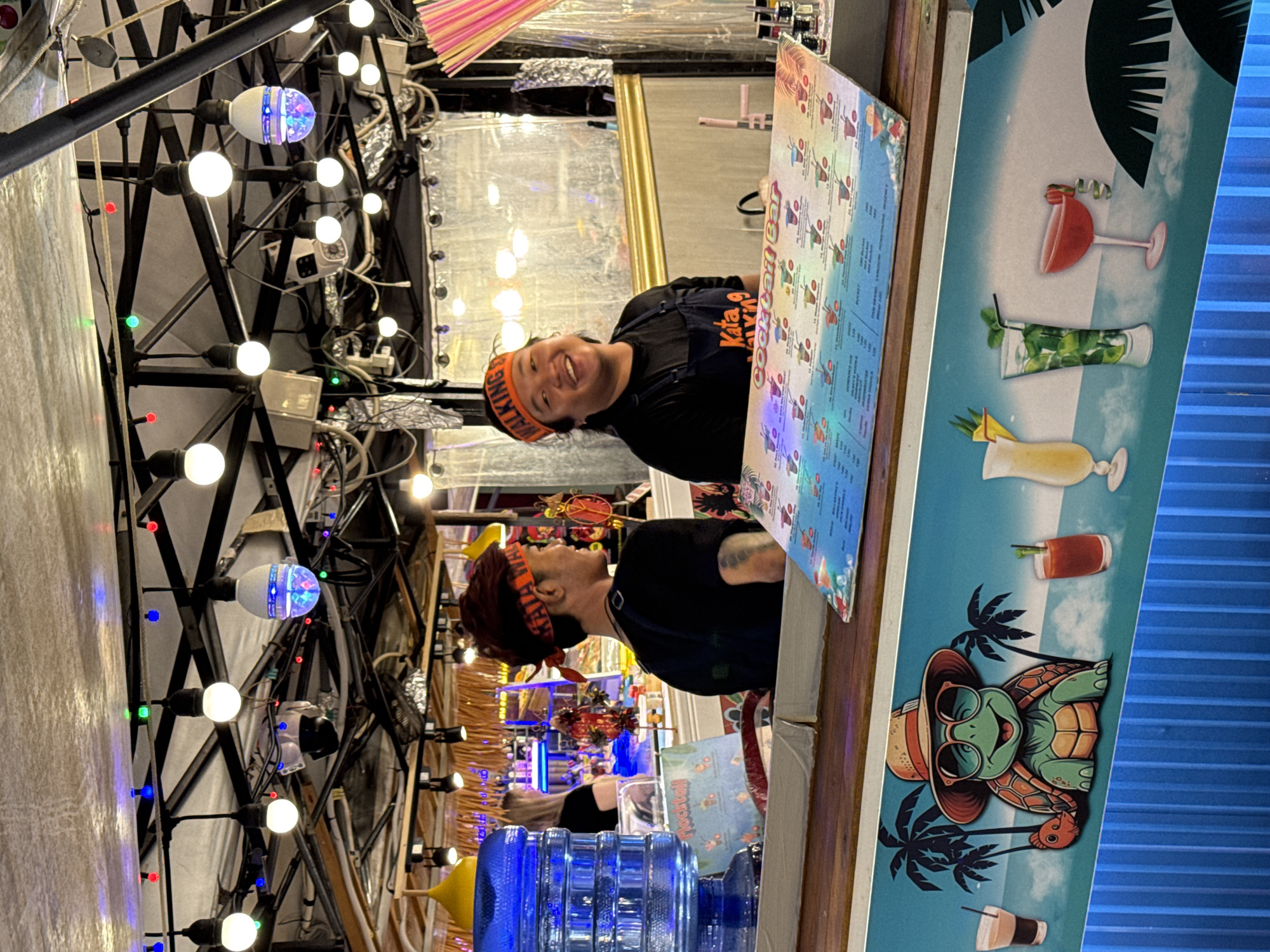 The image depicts an outdoor bar scene, likely in Karon, Thailand, based on the provided information. The bar counter is prominent, with a menu displaying various cocktails. Behind the counter, two individuals are visible. One person, wearing an orange headband and an apron, is smiling and looking towards the camera. The other person, also wearing a headband and an apron, is seen from the side, with their back slightly turned. The background suggests a lively atmosphere with string lights and what appears to be a canopy overhead. A large blue water dispenser is also visible on the left side of the counter. The lower portion of the image features a colorful mural with tropical imagery, including palm trees, a cartoon turtle, and depictions of drinks. The overall scene suggests a casual, tropical bar environment.