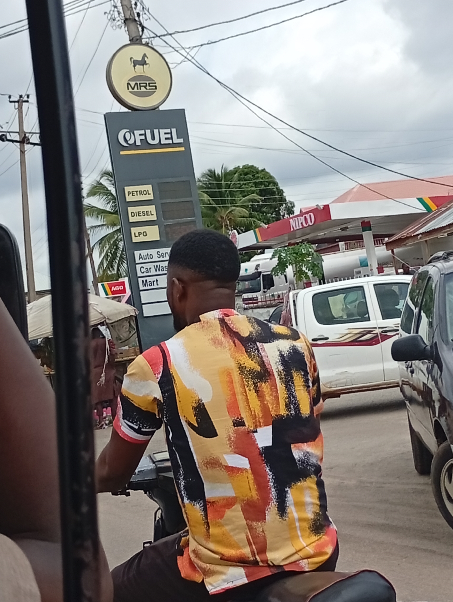 The image depicts a street scene in Lokoja, Nigeria. A man wearing a colorful shirt rides a motorcycle past a gas station branded with the logos of "MRS" and "QFUEL". The gas station sign displays the words "PETROL," "DIESEL," "LPG," "Auto Serv," "Car Wash," and "Mart," suggesting the services offered. In the background, a second gas station with the "NIPCO" logo is visible. Various vehicles, including a white pickup truck and a dark-colored car, are present on the road. Wires overhead contribute to the urban scene. A partially visible stall or shelter is on the left side. The image is taken from the perspective of someone within another vehicle, evidenced by the window frame in the foreground.