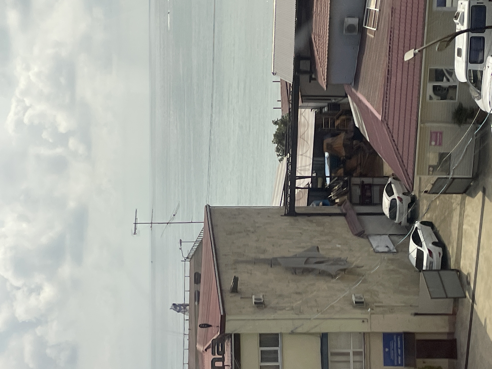 A high-angle, full shot captures an urban scene in Sochi, Russia, with a view of the sea. The foreground features buildings with red-tiled roofs and beige walls. In the middle ground, white cars are parked in an alleyway between the buildings. The background reveals the calm, turquoise sea under a cloudy sky. A few distant boats are visible on the water. The overall impression is one of a coastal city with a mix of residential and commercial buildings.