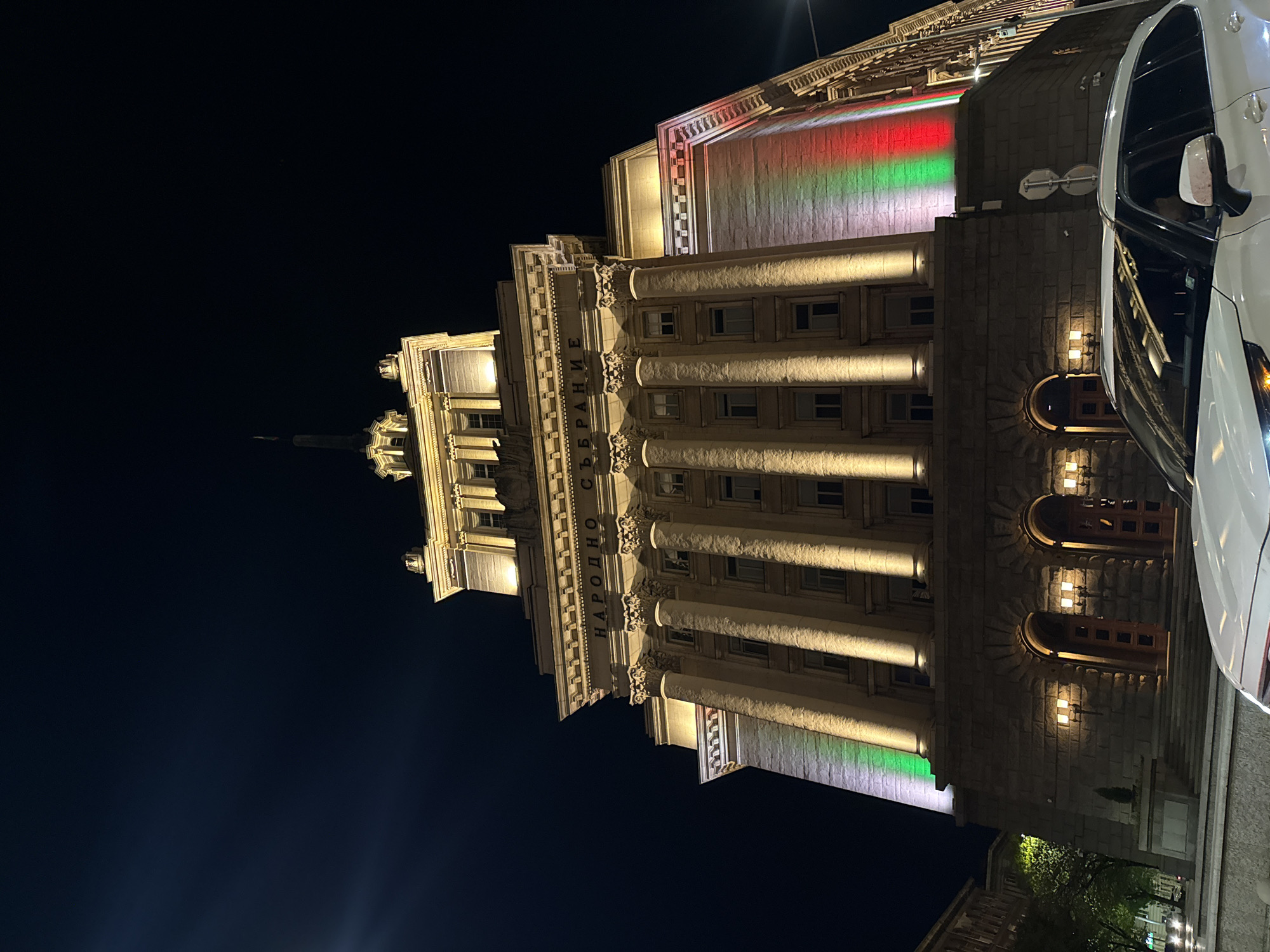 The image depicts the National Assembly of Bulgaria building at night in Sofia, Bulgaria. The building is illuminated with a mix of white spotlights accentuating its neoclassical architectural features such as columns and ornamentation. A section of the building is illuminated in a band of red, white, and green, the colors of the Bulgarian flag. The text "НАРОДНО СЪБРАНИЕ" (National Assembly) is visible on the building's facade. A portion of a white car is visible on the right side of the image. The sky is dark, and minimal street-level details are discernible, aside from some sparse vegetation and distant buildings near the lower right.