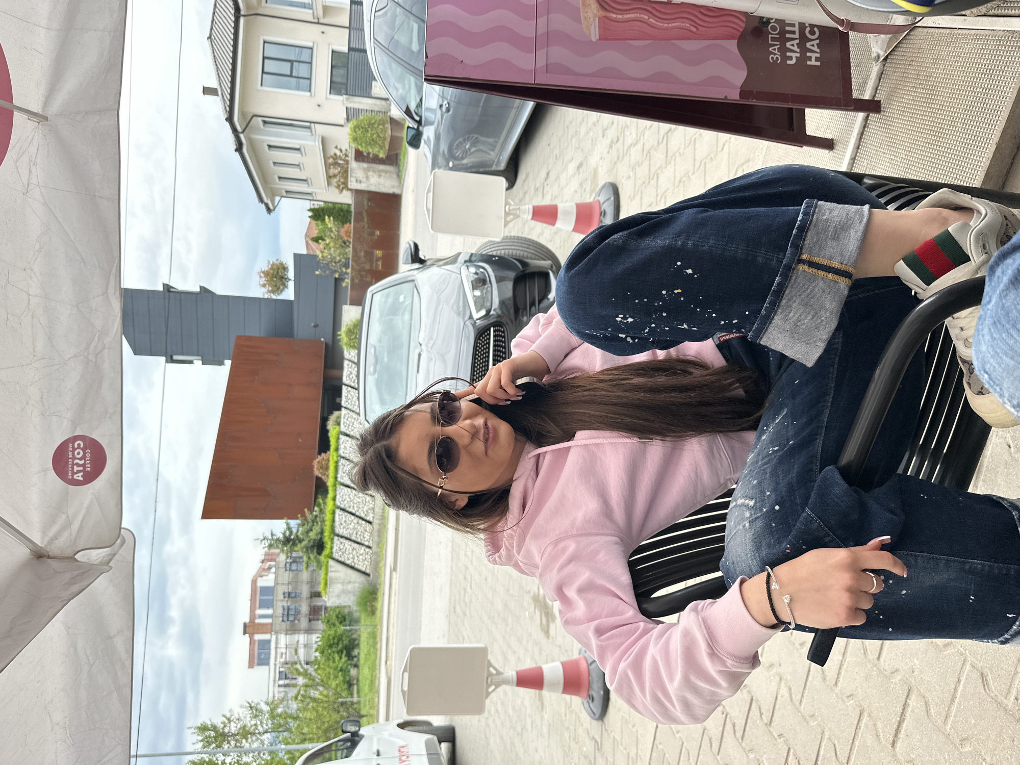 A young woman sits at an outdoor café in Plovdiv, Bulgaria. She wears a pink hooded sweatshirt, paint-splattered jeans with cuffed hems, and Gucci sneakers. She also has sunglasses on and is holding a phone to her ear. The café umbrella is visible on the left, and a sign is partially visible in the top right corner. A grey car is parked nearby, and an orange traffic cone sits on the pavement. Buildings and greenery are visible in the background.