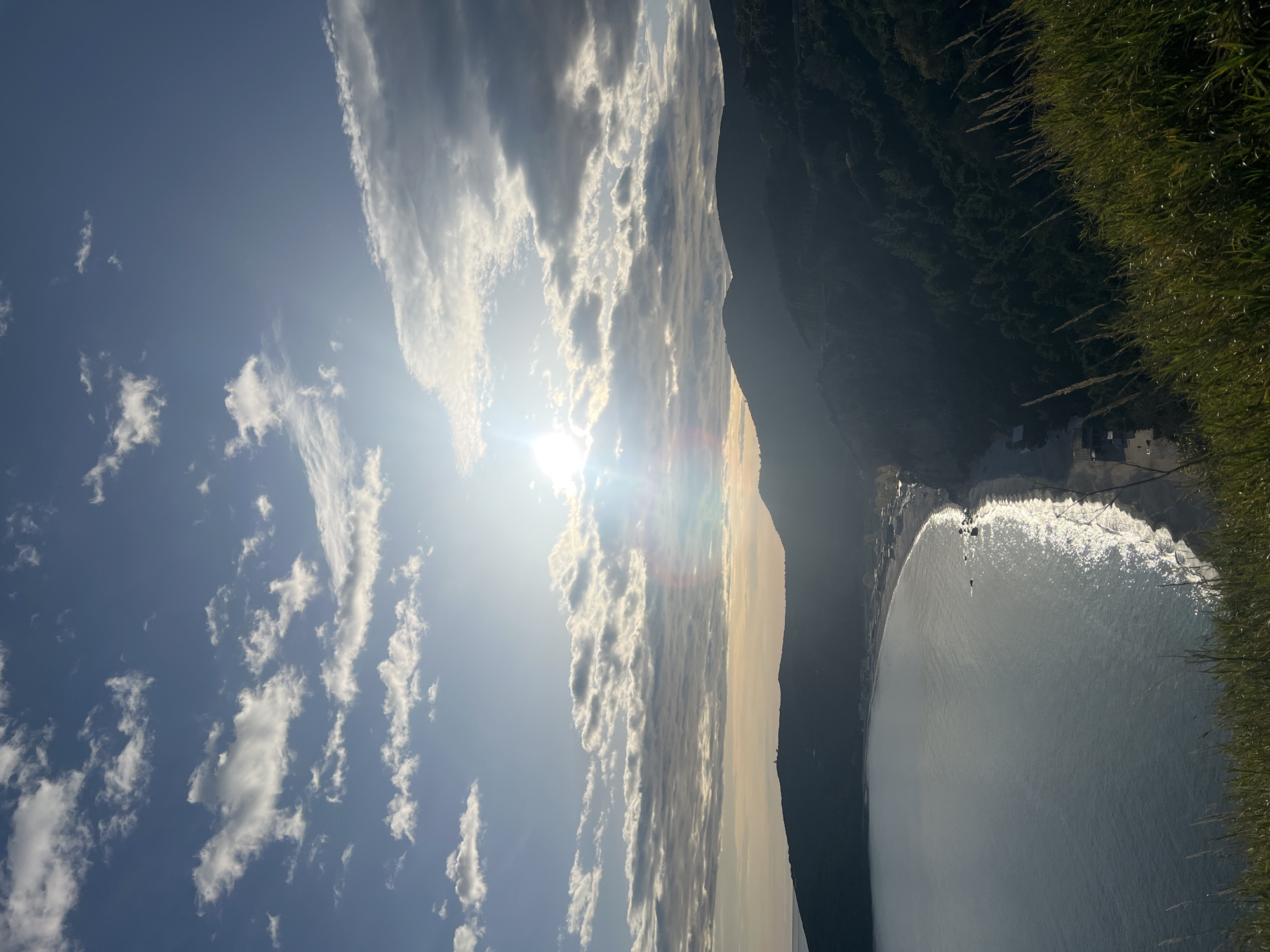 The image is an outdoor shot of a bay in Prijevor, Montenegro. The scene is captured from an elevated perspective, possibly from a cliff or high ground. The sea occupies a large portion of the image, reflecting sunlight, creating a shimmering effect on the water's surface. A sandy beach borders the sea, and a small settlement with buildings is visible along the coast. The right side of the frame shows vegetation, likely grass and shrubs growing on the edge of the cliff. A mountain range dominates the central part of the image, separating the beach area from the sky. The sky is filled with clouds, and the sun is partially obscured by the clouds, causing a bright glare in the sky.