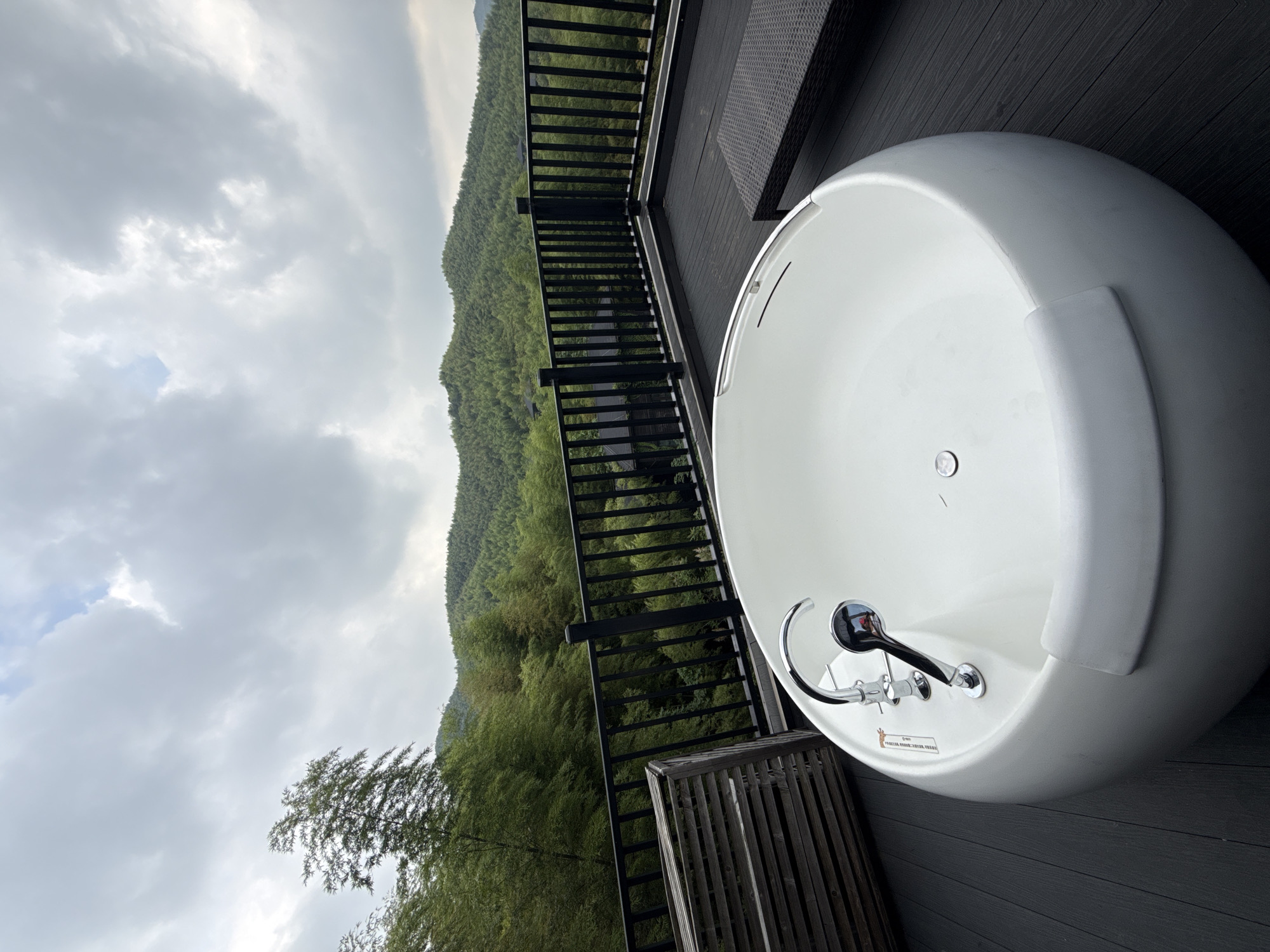 The image depicts an outdoor bathtub on a dark wooden deck, likely a balcony or patio, with a scenic view of lush green mountains and a cloudy sky. The bathtub is white and has a modern, rounded design with chrome fixtures. To the left of the bathtub, there's a wooden privacy screen or partition. In the background, a black railing separates the deck from the natural landscape. Beyond the railing, dense green trees, characteristic of a mountainous or forested area, cover the rolling hills. Several small buildings, possibly part of a resort or residential complex, are visible nestled within the trees. The location is identified as Wuxing District, China, suggesting a tranquil, nature-oriented setting. The overall mood is one of relaxation and serenity, with the bathtub offering a luxurious bathing experience amidst natural beauty.