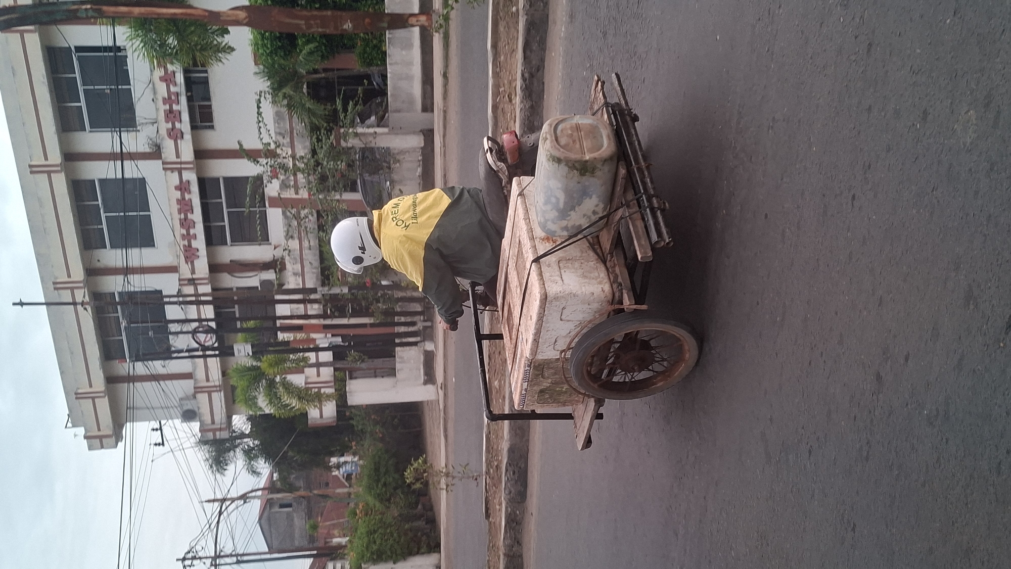 A person wearing a white helmet and a yellow and green jacket is riding a motorcycle with a trailer attached to it. The trailer is carrying two large containers, one white and one grey. The motorcycle and trailer are on a paved road in front of a white building with "WISMA SELA" written in red letters. The building has multiple windows and is surrounded by trees. The sky is overcast. The scene appears to be in Lhokseumawe, Indonesia.