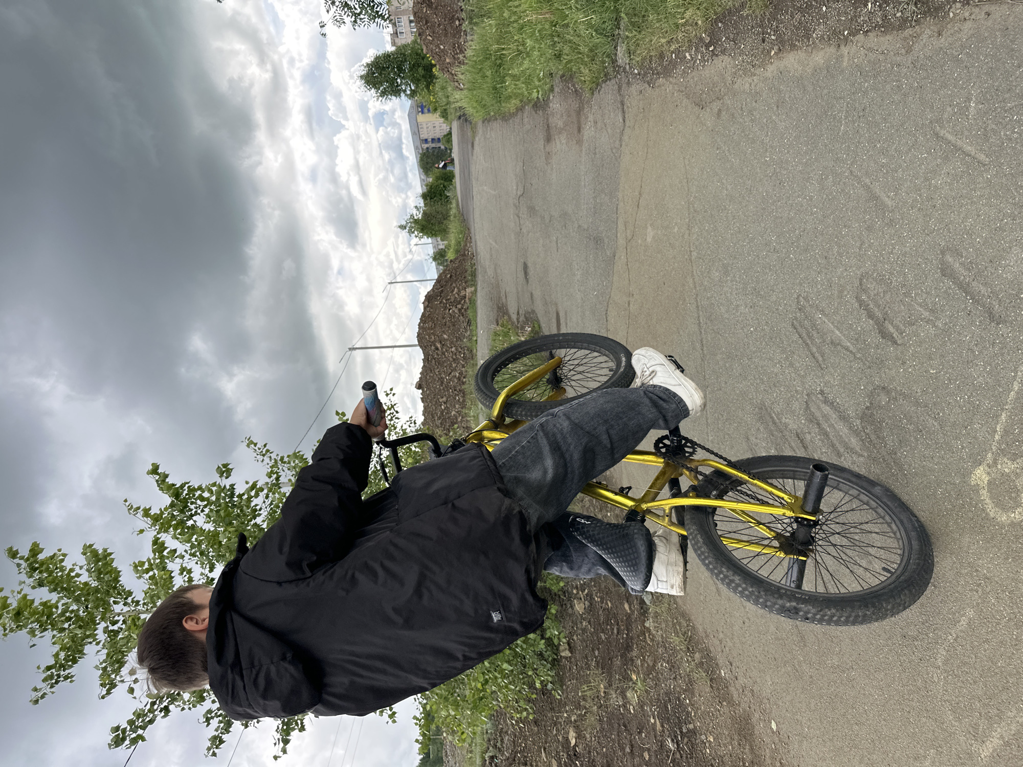 The image depicts an outdoor scene featuring a person on a BMX bicycle under a heavily overcast sky. The person, viewed from behind, is wearing a black jacket, dark grey jeans, and white sneakers, and is straddling or standing next to a metallic golden/yellow BMX bike. A "KV" logo is visible on the lower left side of the black jacket. The bike has black wheels, a black seat, and a blue and white patterned grip on the handlebars; pegs are visible on the rear wheel.
The setting appears to be a paved path or road, possibly an unkempt area or urban fringe, bordered by grass and mounds of dirt or rubble. In the background, there are trees and some distant buildings. The sky is filled with dark, heavy grey clouds, suggesting an overcast day or an approaching storm. Power lines are faintly visible in the mid-ground. The image is oriented sideways, with the natural horizon running vertically through the frame. The time of day is daytime, but muted by the cloud cover. No discernible emotions are visible, as the person's back is to the camera.