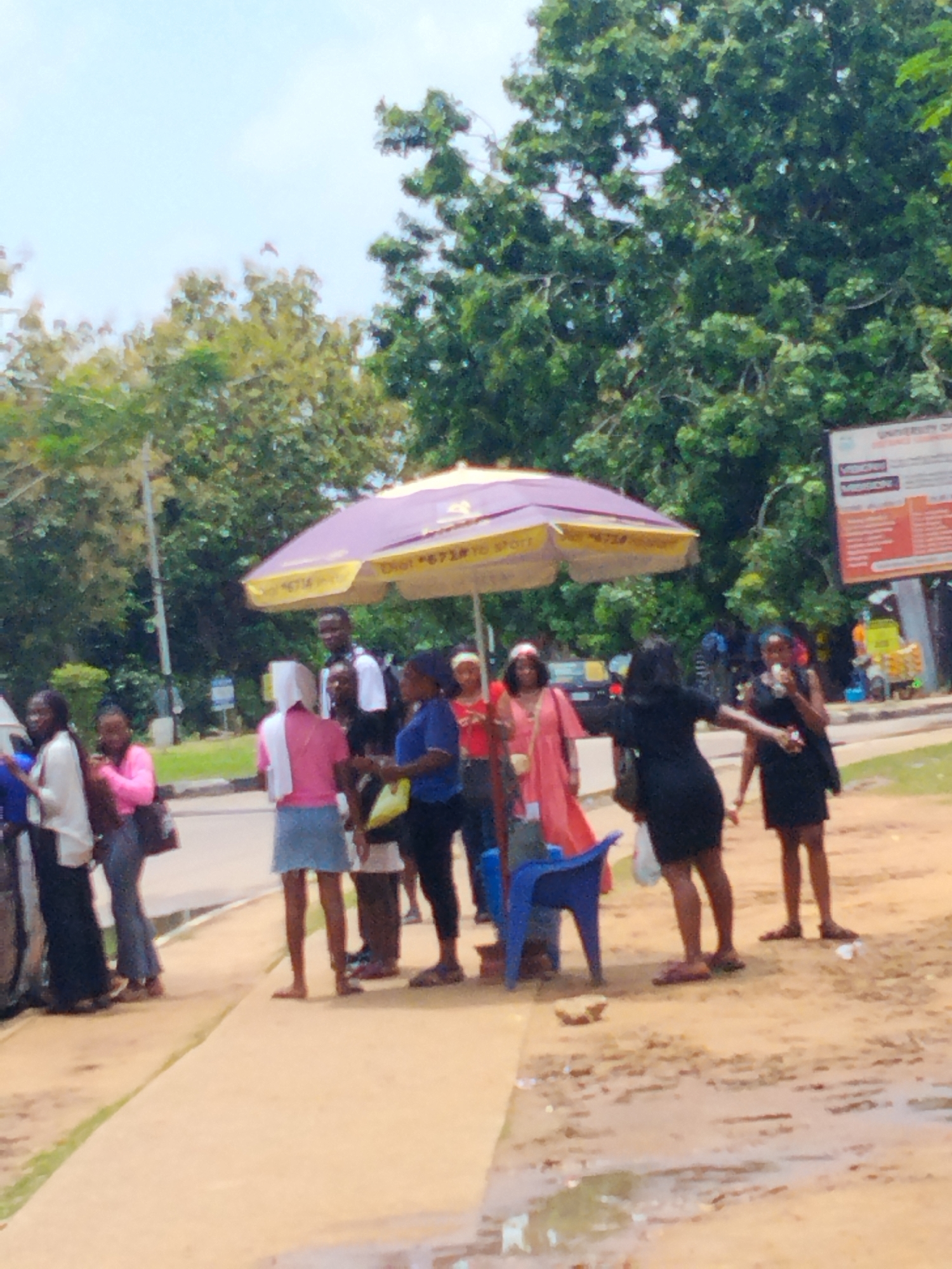 The image depicts a group of people standing on a sidewalk near a street. The environment appears to be a public space in Lagos, Nigeria, as indicated by the “UNIVERSITY OF” sign, a common visual feature in Nigerian cities. The weather seems bright and sunny, with trees providing partial shade.
People: There are around 14 people standing and interacting, all of varying ages and genders. Some are standing under a purple and yellow umbrella.
Subjects: Most of the individuals are standing, some seem to be conversing. Others are eating or holding bags.
Objects: A few of the prominent objects in the scene are an umbrella, a blue plastic chair, the UNIVERSITY OF sign, and a few cars.
Actions: People standing in groups, talking, waiting, walking
Interactions: Individuals are socializing with each other.
Environment: The sidewalk looks like it is made of concrete and adjacent to a dirt or sandy area. A puddle of water is visible on the ground, suggesting recent rain. The background consists of trees and a street with a vehicle parked nearby.