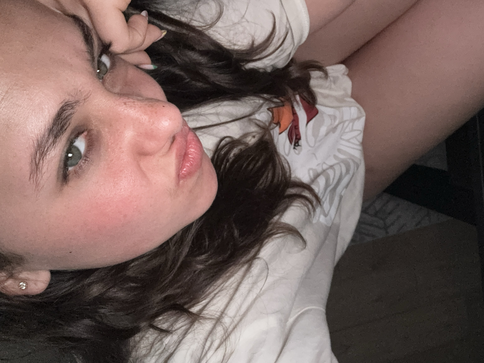 A selfie depicts a woman with light skin, hazel eyes, and dark brown, wavy hair. She is wearing a light-colored shirt with a pattern on the chest. Her lips are slightly pursed, and she is holding her hand near her face. An earring is visible on her left ear. The background is indistinct, but part of a leg and a dark, possibly shadowed area are visible, indicating an indoor setting. There is no visual information to determine the location as Milan, Italy.