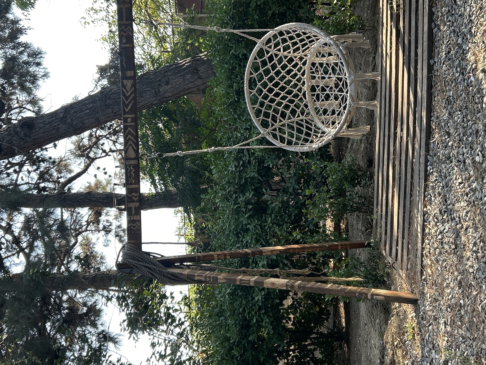 A macrame hanging chair swing is suspended from a wooden beam supported by two wooden posts. The beam is decorated with carved tribal patterns. A thick rope is tied around one of the posts. The swing rests on a wooden platform, which is laid on a ground covered with gravel and pine needles. In the background, there is a dense green hedge and the trunks of tall trees with sparse foliage, suggesting an outdoor, possibly park or garden, setting. The lighting indicates it is daytime. The location is identified as Baku, Azerbaijan. There are no people visible in the image.