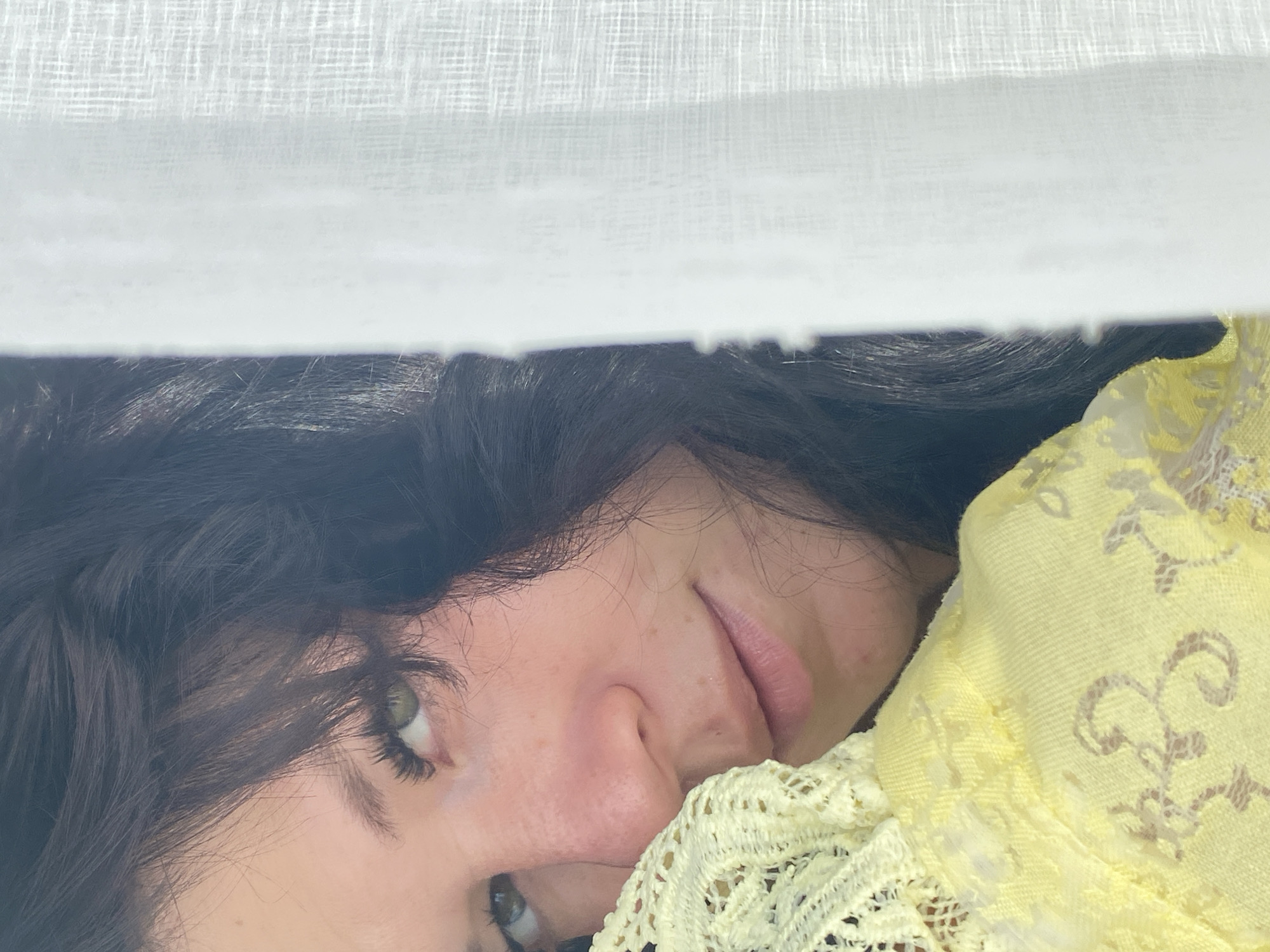 A close-up shot captures a woman's face partially obscured by a white curtain and a yellow garment. She has dark, wavy hair and green eyes, with her left cheek resting on the textured yellow fabric. Her gaze is directed upwards and to her left. The background features a white, translucent curtain, suggesting an indoor setting. The image is well-lit, with soft shadows contributing to a gentle mood.