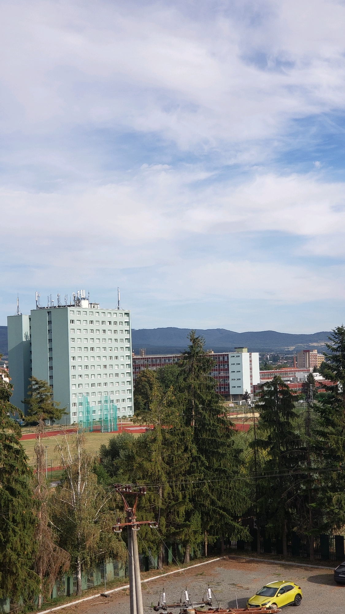 The image depicts a view of the town of Prešov, Slovakia, focusing on an urban landscape with residential and industrial buildings under a partly cloudy sky. In the foreground, a light green, multi-story residential building with numerous windows dominates the left side of the frame. The building is equipped with multiple antennas on its rooftop. Behind it, a long, red-brick industrial or institutional building with white-trimmed windows spans across the mid-ground. Several tall evergreen trees are scattered throughout the scene, with some in the foreground partially obscuring the view of the buildings. A yellow car is parked in a gravel lot in the lower right corner of the image, next to another dark-colored vehicle. The overall environment appears to be a mix of residential areas and infrastructure.