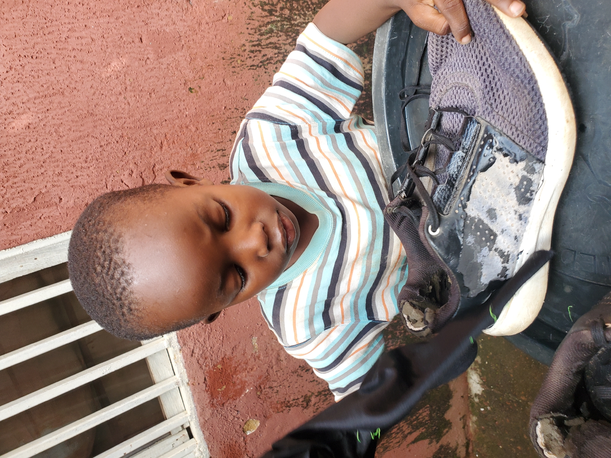 A young Black child, wearing a striped t-shirt, is shown from a medium, slightly high angle in an outdoor setting. The child is looking down intently at a black sneaker they are holding. The sneaker appears wet and possibly dirty. The child's hands are visible, one holding the shoe and the other near it. They are seated in what appears to be a large, black plastic basin or bucket, which is resting on a tire. In the background, there is a textured, reddish-brown wall and a section of a window with white bars. The ground beneath the basin is wet and appears to be concrete with some green patches. This scene is located in the city of Jos, Nigeria.