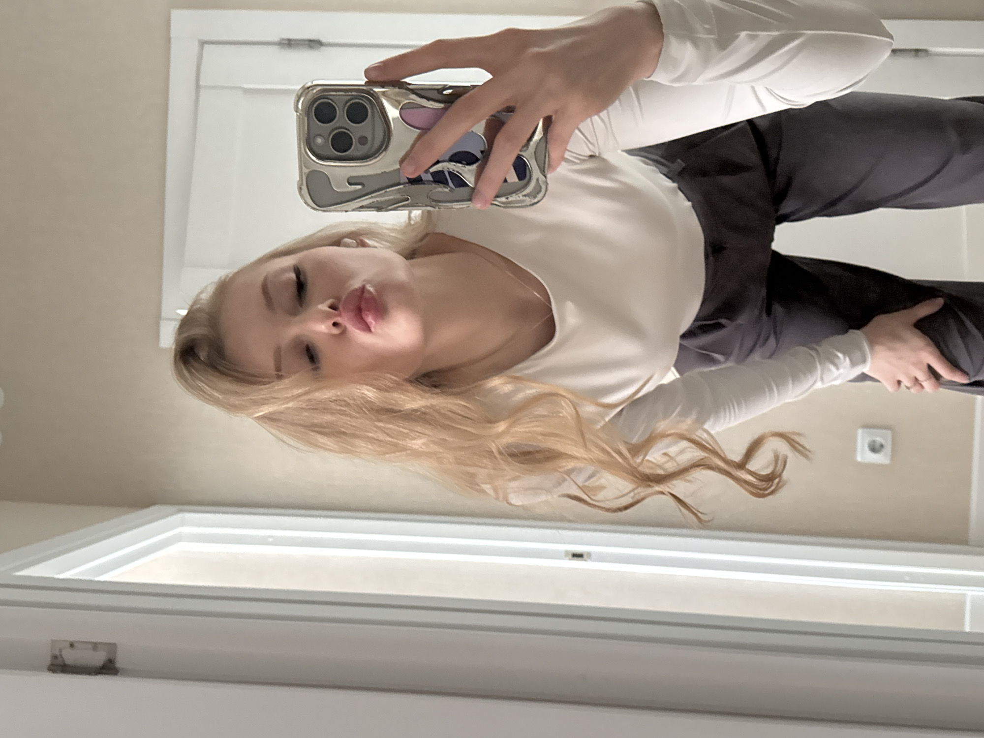 A fair-skinned person with long blonde hair takes a selfie in a mirror. The person has on a white long-sleeved shirt and dark pants. The cell phone in the person's hand has a silver and clear case. The reflection in the mirror shows a room with a door and electrical outlet. The location appears to be a bathroom. This image was taken in Perm, Russia.
