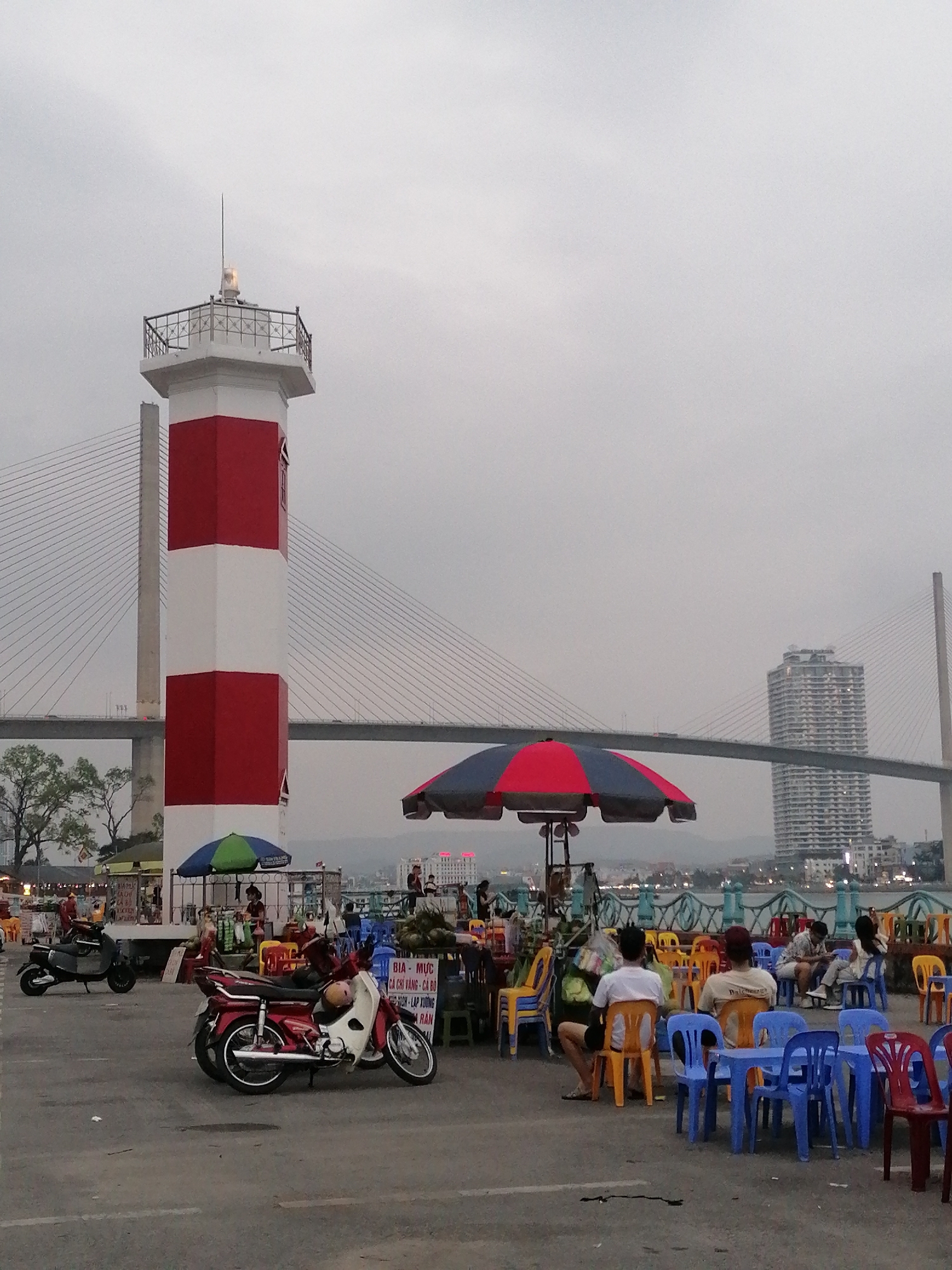 The image depicts a waterfront scene in Bai Chay Ward, Vietnam, under an overcast sky. A red and white striped lighthouse stands prominently on the left side of the frame, with a cable-stayed bridge visible behind it. In the midground, there are clusters of colorful plastic chairs and tables, occupied by several people sitting and interacting. Some of the people are seated and appear to be relaxing or eating. Several motorbikes are parked near the lighthouse. A building can be seen behind the bridge. Parasols offer shade to some of the tables. A food vendor table with signage is also present.