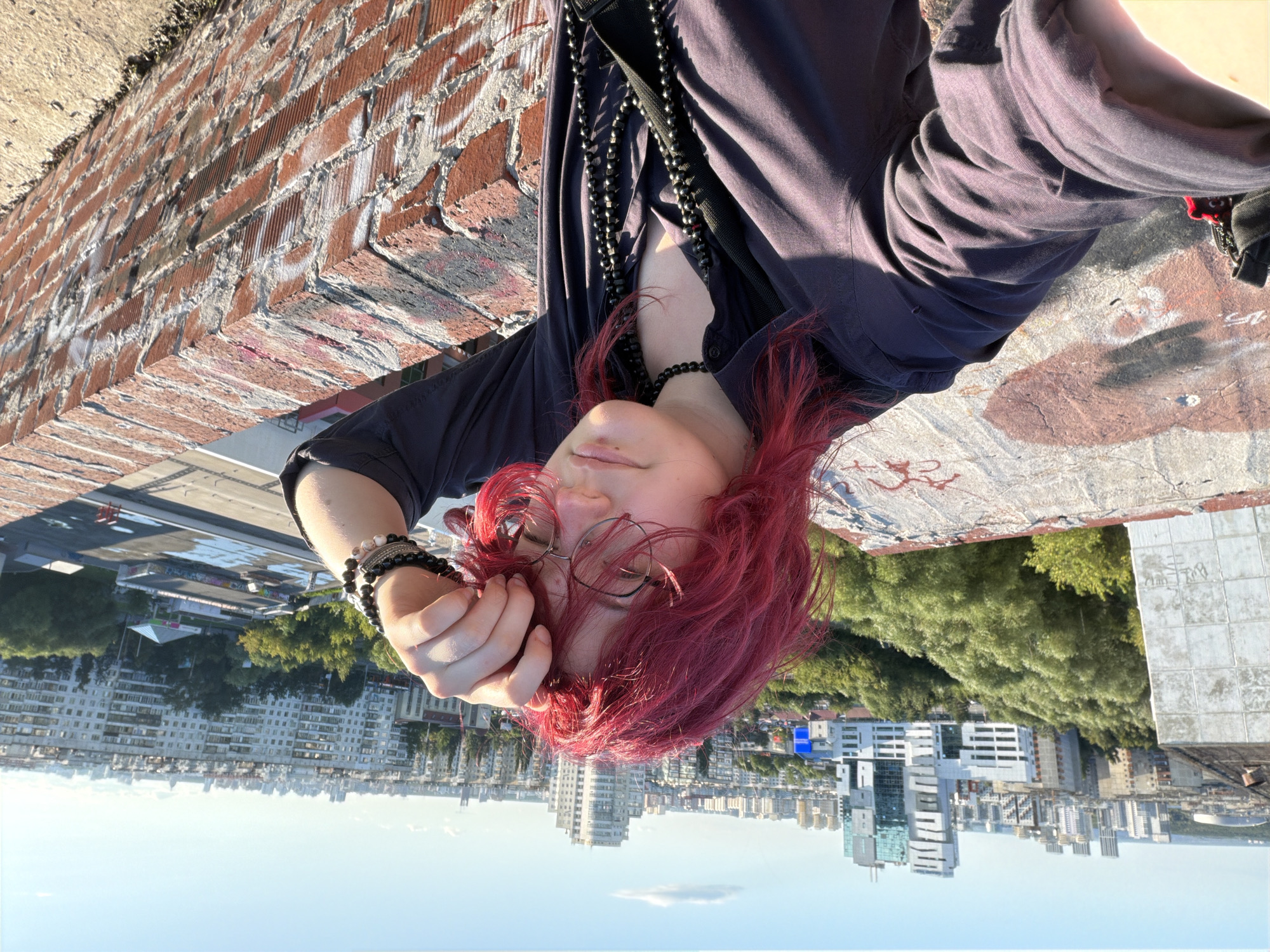 A person with vibrant red, messy hair and glasses is taking a selfie on a rooftop overlooking a city. They are wearing a dark-colored collared shirt, a choker necklace, and several long beaded necklaces. A black strap from a bag is visible across their chest. The person is touching their forehead with their right hand. The background shows a cityscape with various buildings, including modern high-rises and apartment blocks, under a clear blue sky. The rooftop itself is made of concrete and brick, with some graffiti visible on the concrete surface. The location is identified as Chelyabinsk, Russia.