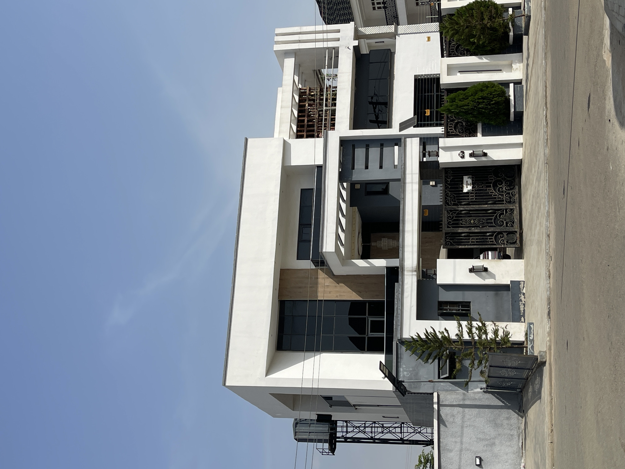 The image displays a modern, multi-story building under a bright, clear blue sky in Gwarinpa, Nigeria. The architecture is contemporary, featuring a striking contrast of white exterior walls with dark grey and black accents.
The building showcases a clean, geometric design with multiple levels and prominent large, dark-tinted windows. Several balconies are visible, some with white railings and internal wooden paneling. A large, ornate black metal gate serves as the main entrance to the property, flanked by exterior wall sconces. Smaller black metal gates and fences are also present around the perimeter.
Greenery is incorporated into the design with several small, well-maintained evergreen trees or shrubs planted along the building's base. To the left, a water tank is supported by a metal structure. On the lower right, near a smaller dark gate, several dark panels, possibly solar, are installed at ground level. A paved road or driveway runs alongside the building in the foreground.
The scene is set on a bright, sunny day, indicated by the vibrant blue sky and visible shadows. There are no people or discernible activities taking place. A small, white sign with unreadable black text is affixed to the main black metal gate.