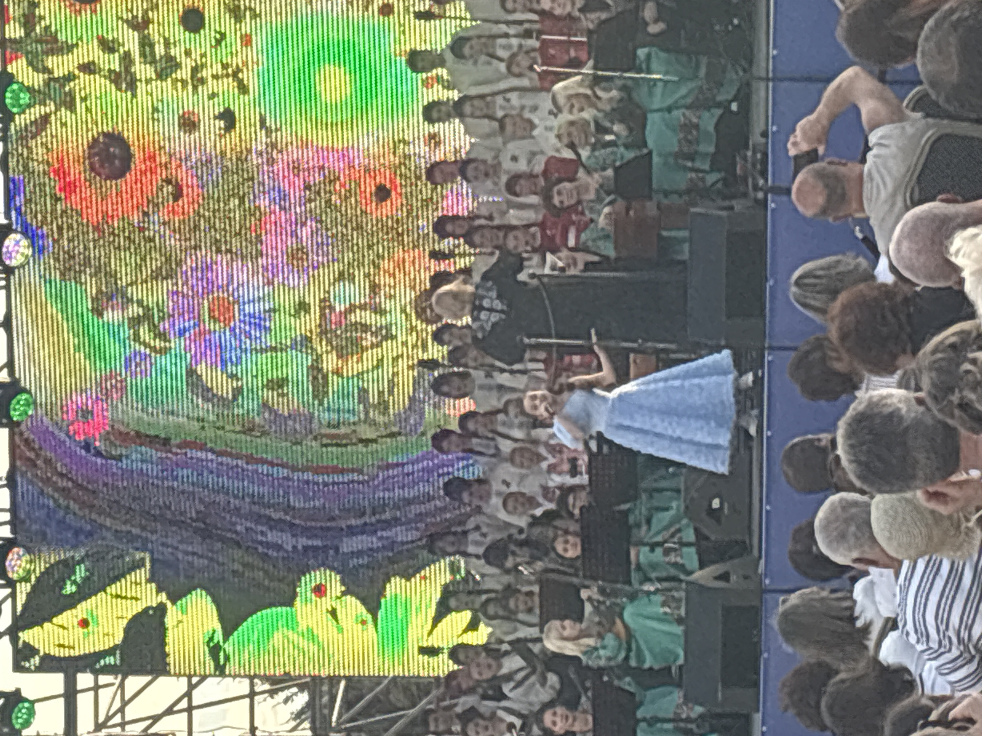 A crowd is gathered to watch a performance on a stage. In the foreground, people are seated and standing, some holding phones to take pictures. The stage is set up with a large screen displaying a colorful, abstract floral pattern. A group of performers, including a soloist in a light blue dress and a conductor in a dark, ornate robe, are on stage. The soloist is singing into a microphone, and the conductor appears to be directing the other performers, who are arranged behind them, some holding sheet music. Several large speakers and lighting equipment are visible on and around the stage, suggesting a formal concert or festival event. The overall atmosphere appears to be festive and engaged. The event is taking place in Astrakhan, Russia.