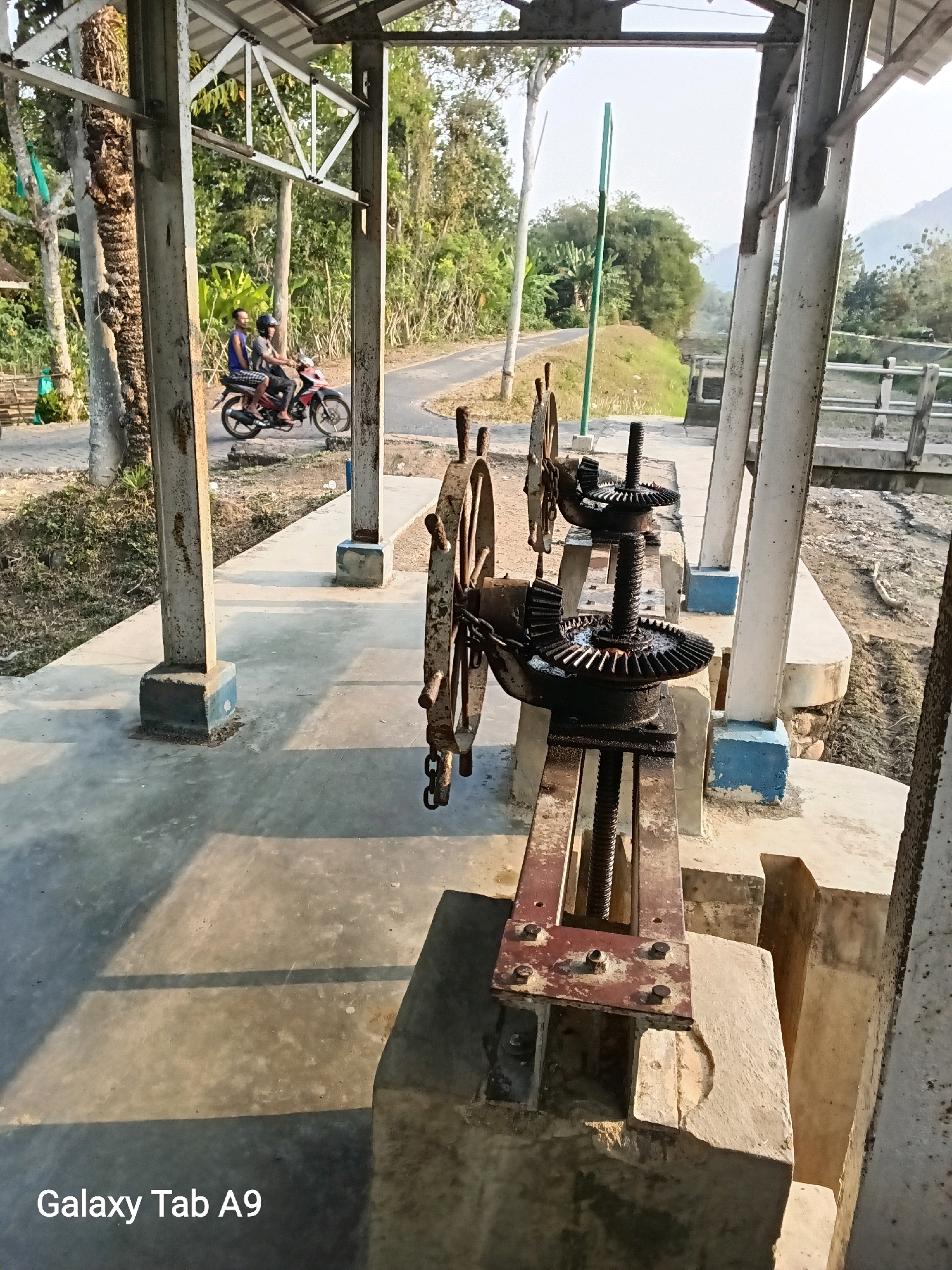 The image captures an outdoor scene in Sumberingin, Indonesia, focusing on a mechanical water gate system. In the foreground, two prominent, weathered gears are attached to a threaded rod, part of the water gate's lifting mechanism. These gears are rusty and show signs of heavy use, suggesting an industrial or agricultural setting.
Behind the water gate mechanism, a concrete pathway leads away from the viewer. On this pathway, two individuals are riding a motorcycle. The person in front is driving, while the passenger sits behind. They are moving away from the camera, towards a rural landscape. The road curves to the left, lined by lush green vegetation and trees.
The scene is framed by a structure with white metal support beams and a corrugated roof, providing shade over the pathway. The background reveals a gentle slope of land covered in greenery, with distant hills or mountains under a hazy sky. The overall impression is one of a functional, utilitarian environment integrated into a natural, possibly rural, setting. The lighting suggests it is daytime, with shadows cast on the concrete by the overhead structure. The bottom of the image contains a watermark "Galaxy Tab A9".