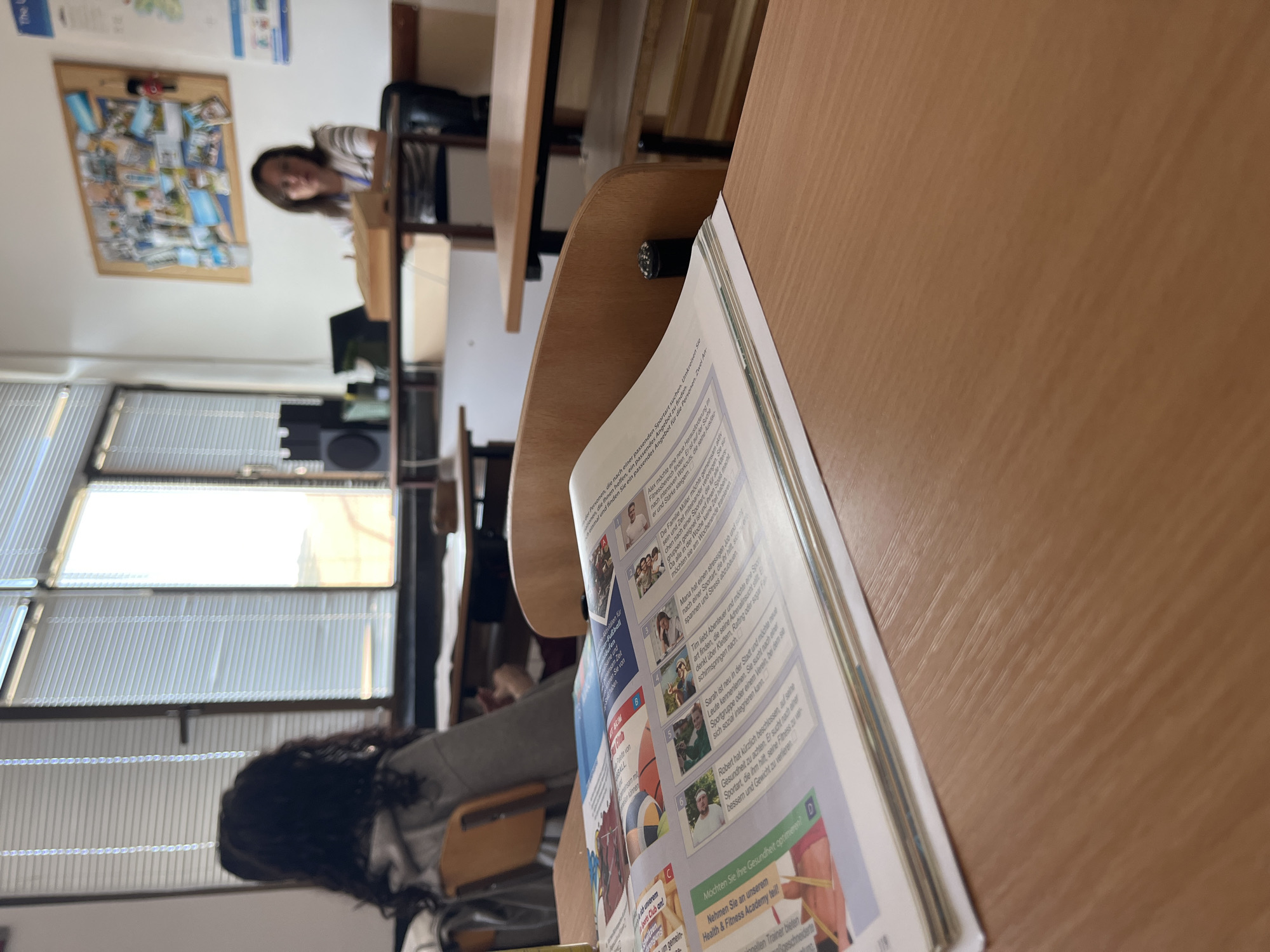 A classroom scene is depicted, with a student seated at a desk in the foreground, facing away from the camera and towards the windows. The windows are covered with blinds, and sunlight streams through, creating bright areas. A magazine or textbook lies open on the desk in front of the student. In the background, another student is seated at a desk, facing a whiteboard. A teacher or instructor is also visible in the background, sitting at a desk. A bulletin board with various pictures and documents is displayed on the wall. The room appears to be a typical classroom setting with desks and chairs arranged in rows. The location is identified as Zlatograd, Bulgaria.