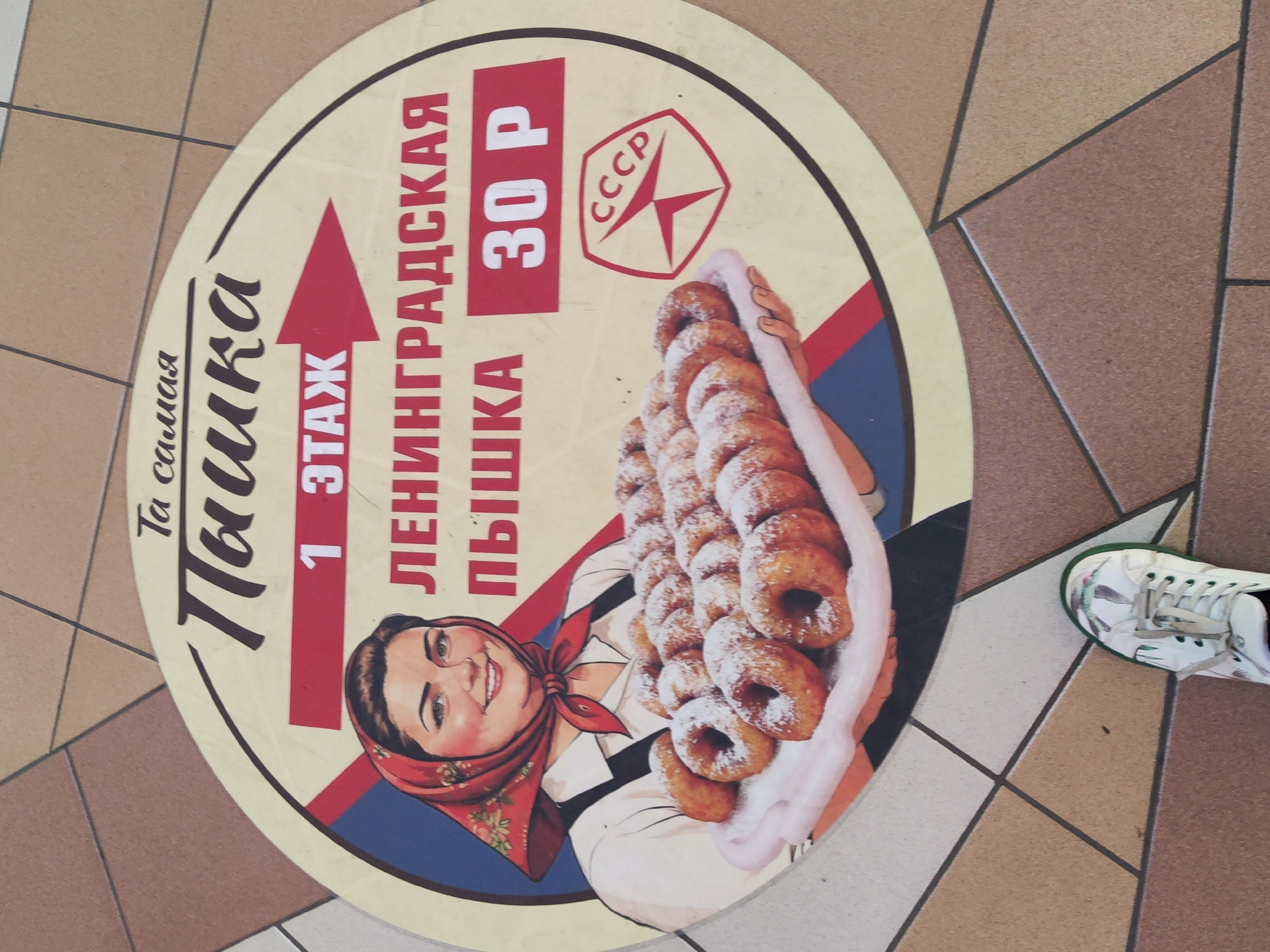 The image shows a round floor advertisement for a "Pyshka" shop, a local Saint Petersburg donut. The advertisement is printed on a surface placed on a tile floor. The ad features the words "Самая Пышка" in Russian, "1 этаж" (1st floor) with a red arrow pointing upward, and "Ленинградская Пышка" (Leningrad Pyshka). It also indicates a price of "30 P" (30 Rubles). The design includes the CCCP logo and an image of a woman holding a tray of sugar-dusted donuts. A partial view of a white sneaker is visible in the lower right corner of the frame. The environment consists of a tile floor, likely in a shopping mall or commercial building in St. Petersburg, Russia.