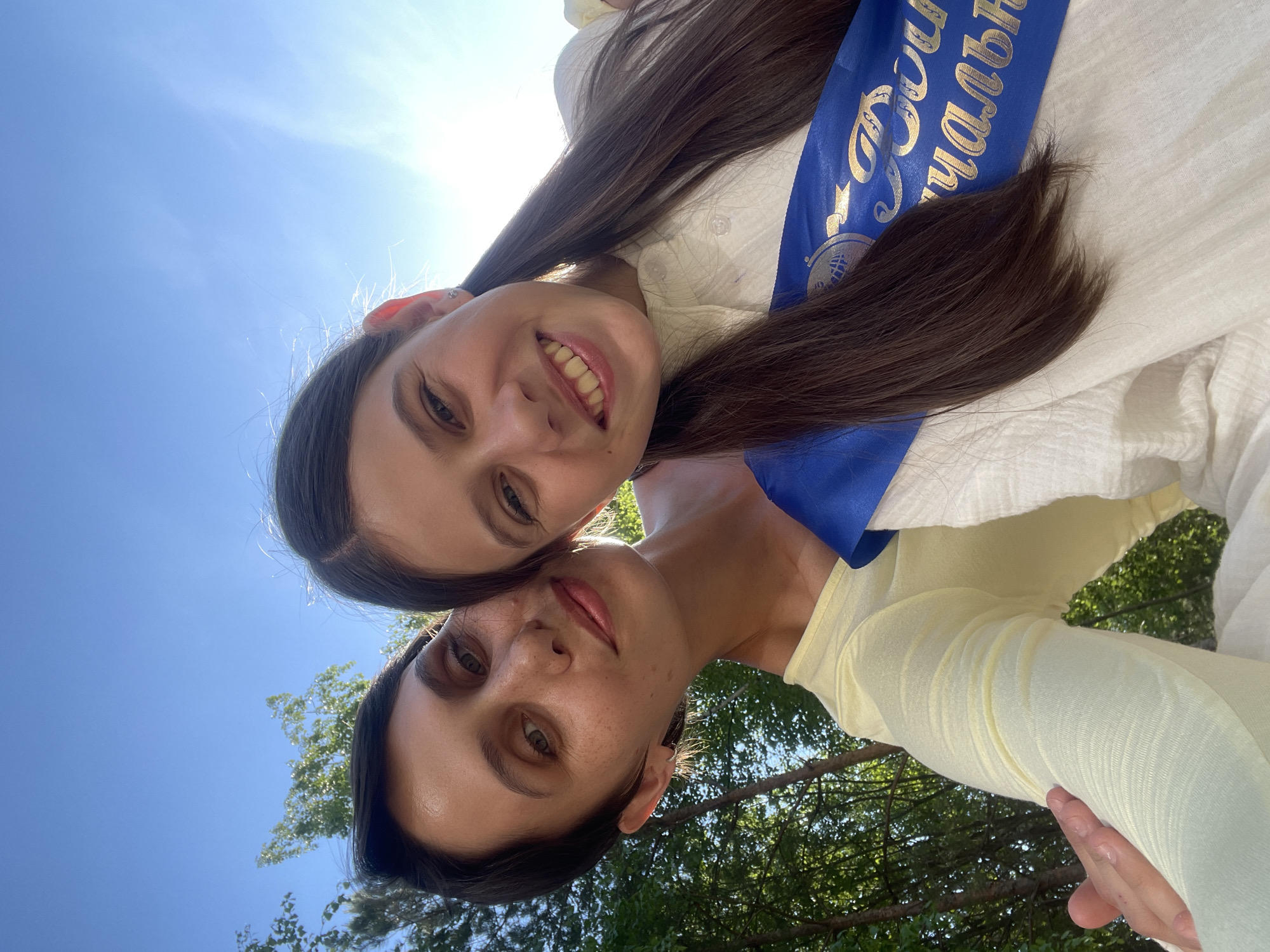 Two young women are pictured outdoors on a sunny day. The image is oriented at an angle, showing the sky and trees in the background. The top woman has long, brown hair and wears a white shirt with a blue sash bearing gold lettering. The bottom woman wears a yellow shirt. They appear to be close together, possibly taking a selfie. The location is possibly Izhevsk, Russia, based on the writing on the sash.