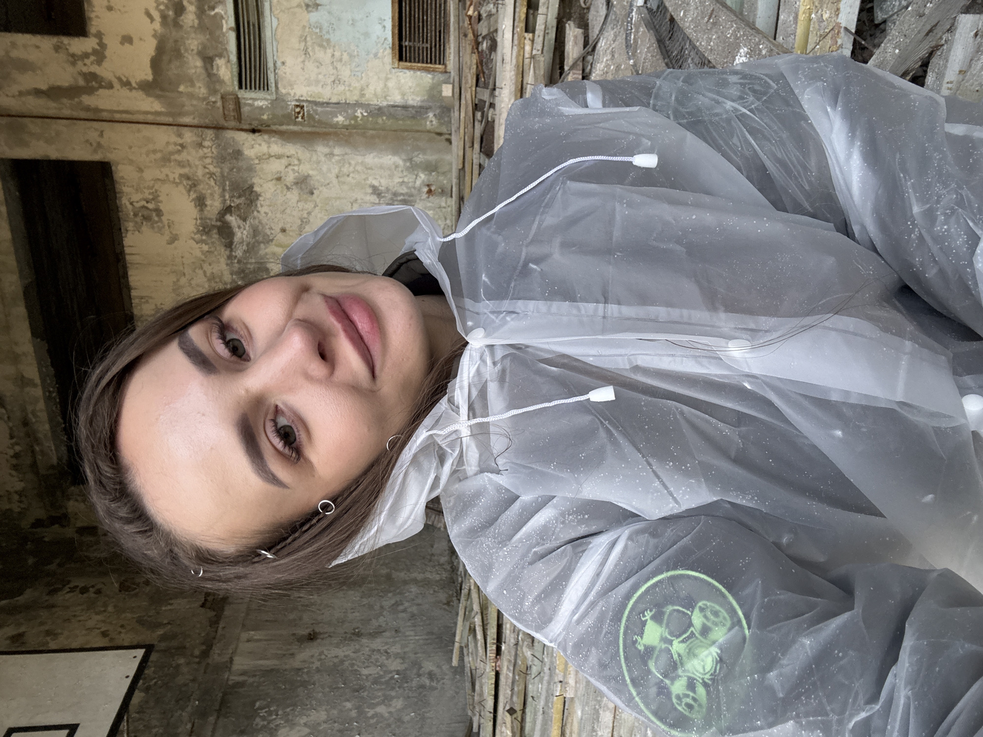 A woman wearing a clear plastic poncho is in an interior location within Prypiat, Ukraine. The backdrop features decaying walls and structures. The plastic poncho has a green logo on the lower portion.