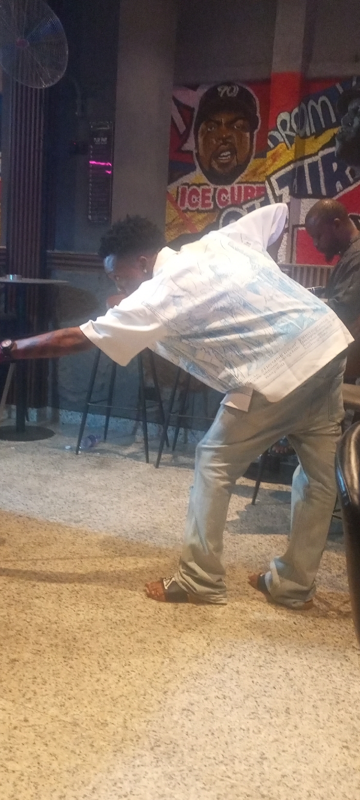 An indoor photograph captures a man deeply focused on a game of billiards, likely in a bar or recreational space in Lokoja, Nigeria. The main subject, a dark-skinned man with short, dark curly hair, is seen from a side-rear angle, bending low with his right arm extended forward, indicating he is about to make a shot. He is wearing a white short-sleeved button-up shirt with light blue, unreadable patterned graphics, light blue jeans, and dark sandals with a distinct white 'V' logo on the strap.
In the background, on the right, another dark-skinned man wearing a dark shirt is partially visible. The environment features a light-colored, speckled floor and dark walls. A prominent, colorful mural covers the back wall, displaying a caricature of rapper Ice Cube wearing a baseball cap with what appears to be "WC" or "YO" on it. Below his image, the text "ICE CUBE" is clearly visible. To the right of Ice Cube, fragmented words like "PROM" or "FROM" and "TRE" are part of the mural's design. An industrial-style fan is mounted on the upper left wall, and dark bar stools are scattered in the mid-ground. A light fixture with a pink glow is also visible on the back wall. The lighting suggests an evening or nighttime setting. The man's posture conveys intense concentration on his game.
