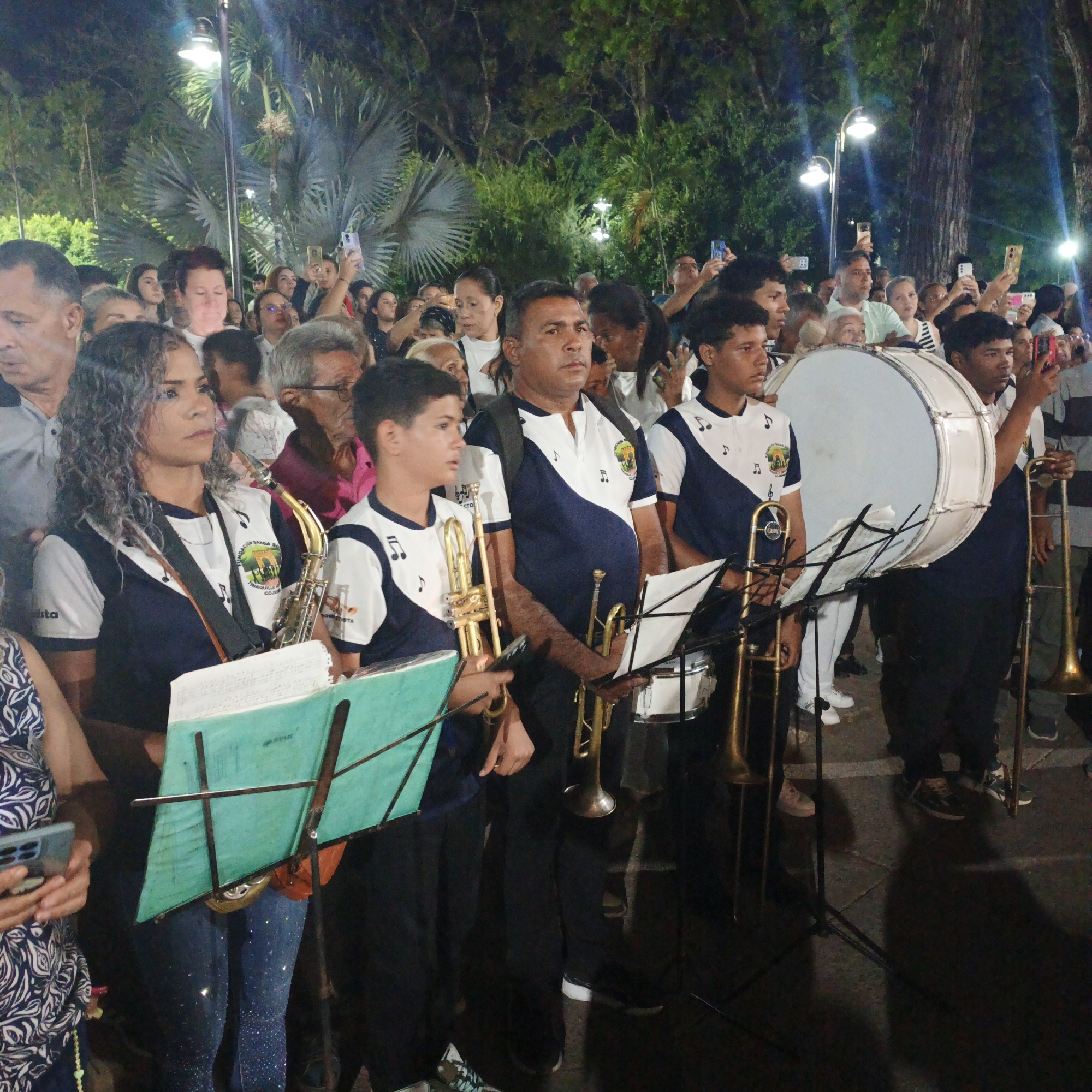 The image depicts a musical band performing in a public square, presumably in the city of Tinaquillo, Venezuela. The band members, wearing matching white shirts with blue accents, are positioned in the foreground. Each musician holds a brass instrument, and some have music stands with sheet music. Behind the band, a crowd of people stands, many of whom are holding up smartphones to record the performance. The environment includes trees, street lights, and a paved area underfoot. It is nighttime.