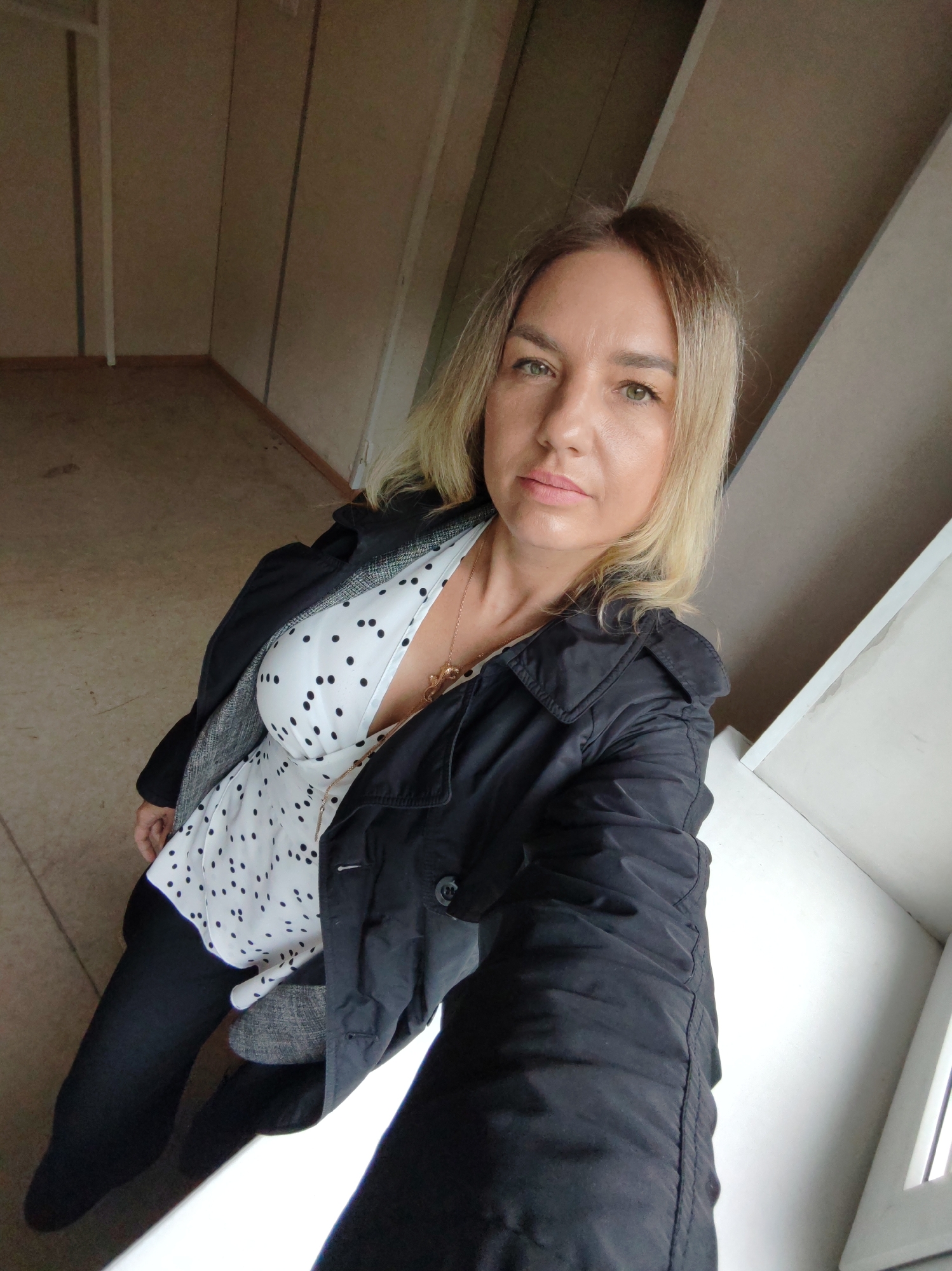 A woman with blonde hair and light-colored eyes is featured in a selfie taken indoors. She looks directly at the camera with a neutral to slightly contemplative expression. She is wearing a black jacket layered over a white top with black polka dots, and a gold-toned necklace with a pendant. Her dark trousers or leggings are partially visible towards the bottom of the frame.
The background shows an interior space, likely a room with light beige or light brown paneled walls, indicated by vertical lines. The floor appears to be a similar light shade, possibly carpeted or linoleum. A white structure, potentially a window frame or part of the wall, is visible in the lower right portion of the image. The overall setting appears functional or minimally furnished. There are no specific visual cues within the image to determine the time of day or weather. No visible text is present. The provided context indicates the location as Petropavlovsk-Kamchatsky, Russia.