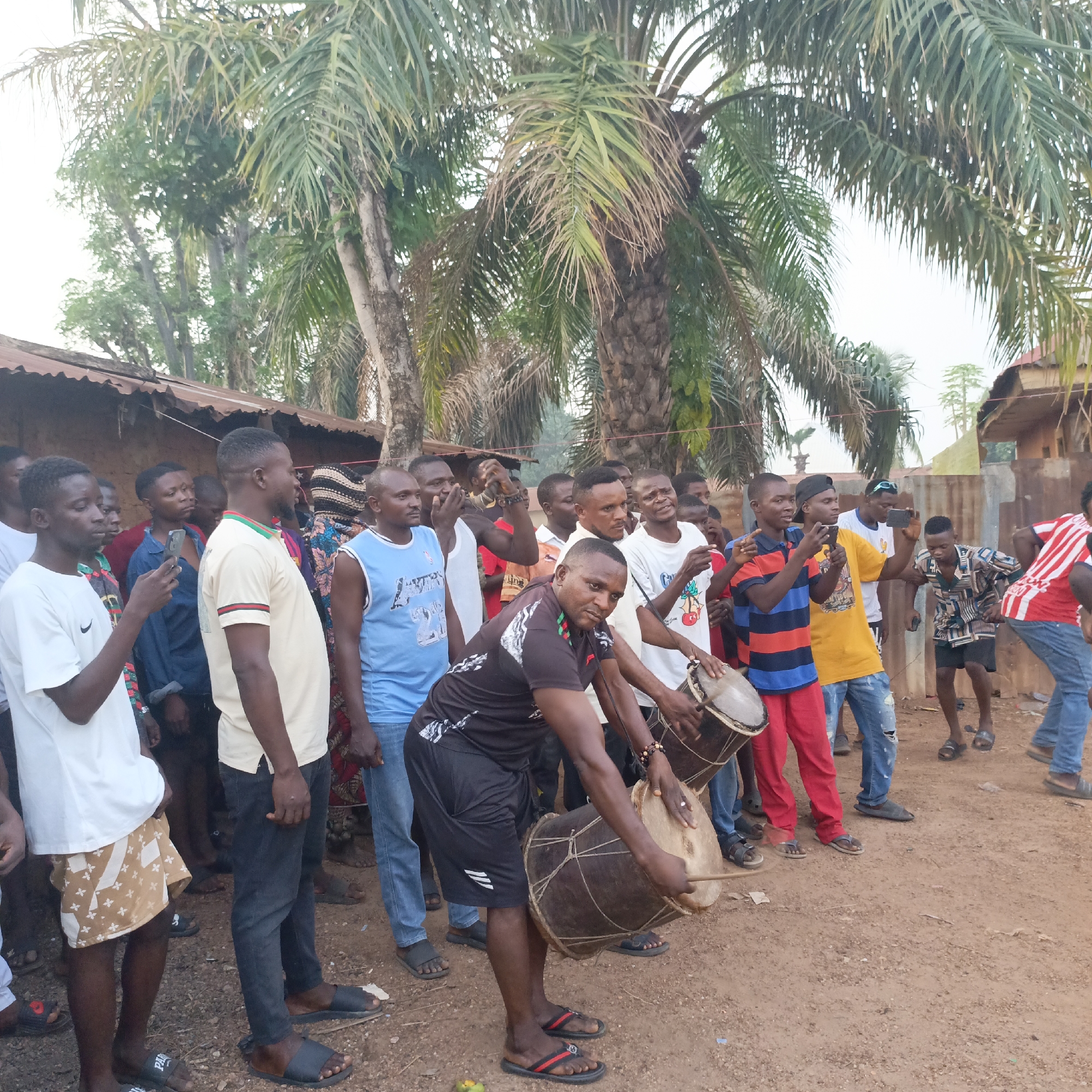 A group of men are gathered in an outdoor location in Akwanga, Nigeria. The men are of African descent and are dressed in casual clothing, with some wearing sandals. A man in the foreground is playing a large drum, while others observe and some record with their mobile phones. A palm tree rises behind the crowd, and a building with a corrugated metal roof is visible in the background, indicating a residential area. The ground is dirt, suggesting an unpaved street or yard.