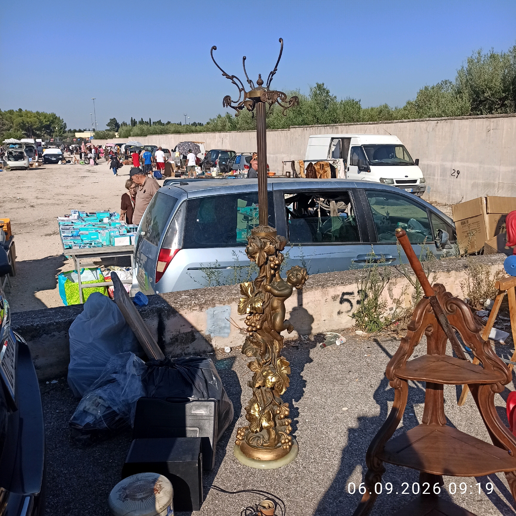 This image depicts an outdoor market, likely a flea market or a car boot sale, on a sunny day. The scene is located in Sannicandro di Bari, Italy, as indicated by the timestamp. In the foreground, a decorative gold-colored coat rack with a cherubic figure and grapevines is prominently displayed. To the right of the coat rack, a vintage wooden corner shelf unit is visible. Further back, a light blue van is parked, and beyond that, various cars, stalls, and people are spread across a dusty, gravelly ground. The background features a concrete wall and trees, suggesting a semi-urban or suburban setting. Several people are browsing or interacting at the stalls. Bags of trash are piled near the foreground. The overall atmosphere suggests a busy market day.