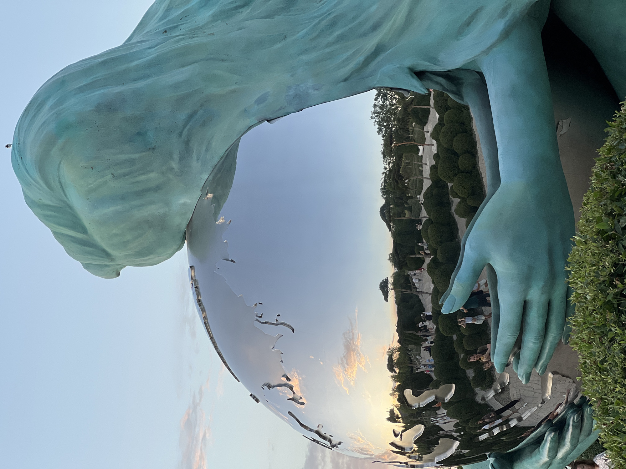 A close-up, rotated shot captures a large, teal-colored sculpture with smooth, flowing lines, reminiscent of draped fabric or a human form. The sculpture features large hands gently cradling a reflective, globe-like object. The surface of this globe is highly polished, mirroring the surrounding environment.
Reflected in the globe are several elements: a clear sky with soft, pastel-colored clouds, suggesting either sunrise or sunset. Below the sky, a manicured garden is visible, featuring neatly trimmed bushes, trees, and paved pathways. Several people are scattered throughout the garden, appearing small in the reflection, and seem to be enjoying a leisurely stroll or gathering. The overall atmosphere conveyed by the reflection is one of calm and pleasant outdoor activity.
The sculpture itself has a patina that suggests it may be made of bronze or a similar metal, weathered to achieve its distinct teal hue. The detailed rendering of the hands, with defined fingers and knuckles, adds a sense of realism to the artistic piece. The globe, with its smooth, unbroken surface, acts as a dynamic mirror, distorting and re-presenting the world around it. The composition, with the sculpture dominating the foreground and the reflected scene occupying a significant portion of the frame, creates a visually engaging juxtaposition of art and environment.
The lighting suggests it is late afternoon or early evening, given the warm tones in the sky and the long shadows that might be present but are not explicitly visible. The setting appears to be an open public space, likely a park or garden designed to showcase sculptures, given the formal arrangement of the landscaping. No text is visible in the image.